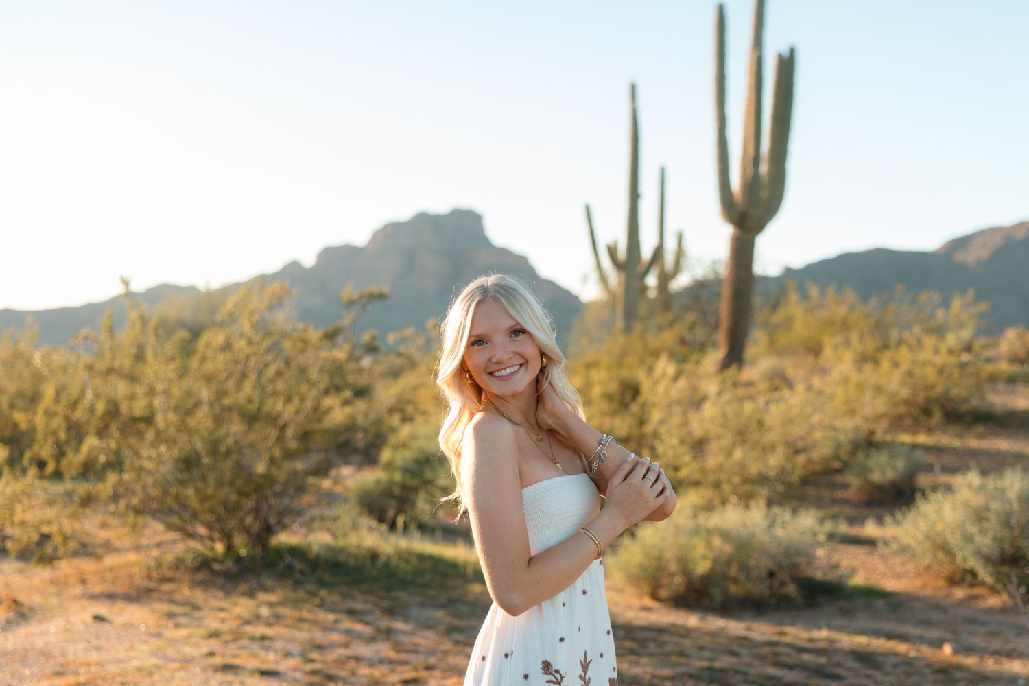 Blonde woman in white strapless dress smiling in sunny Arizona desert with saguaro cactus backdrop.