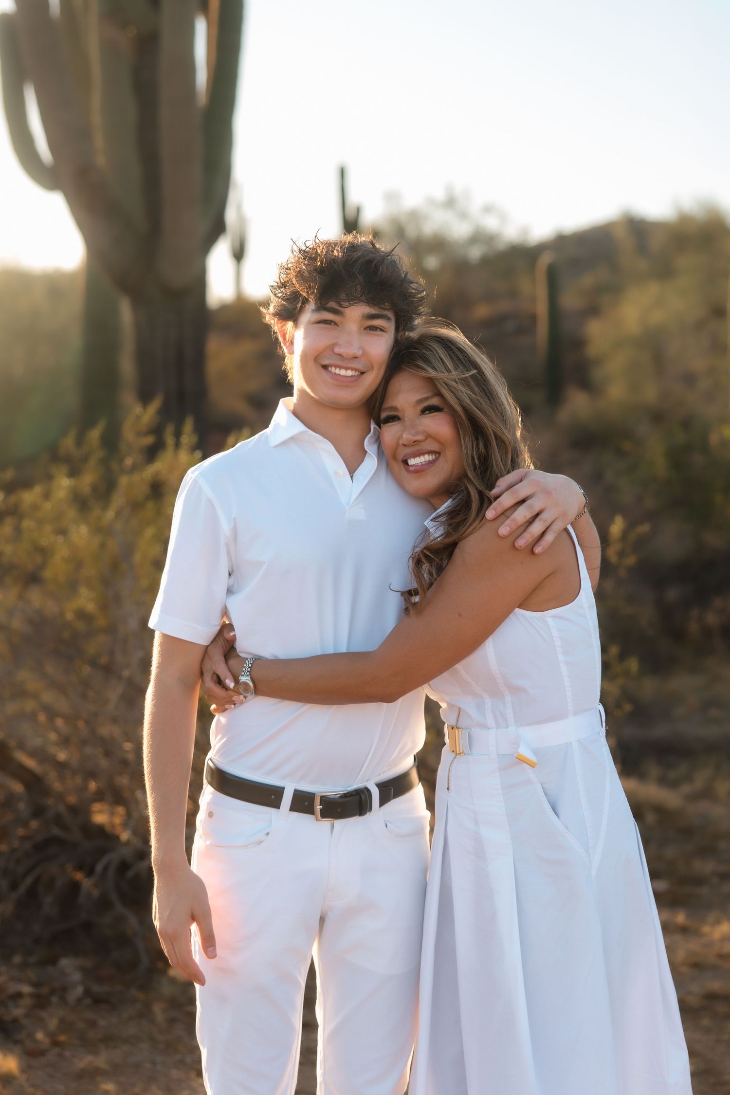 Teenage boy and woman in white outfits hugging warmly in desert with saguaro cactus at sunset.