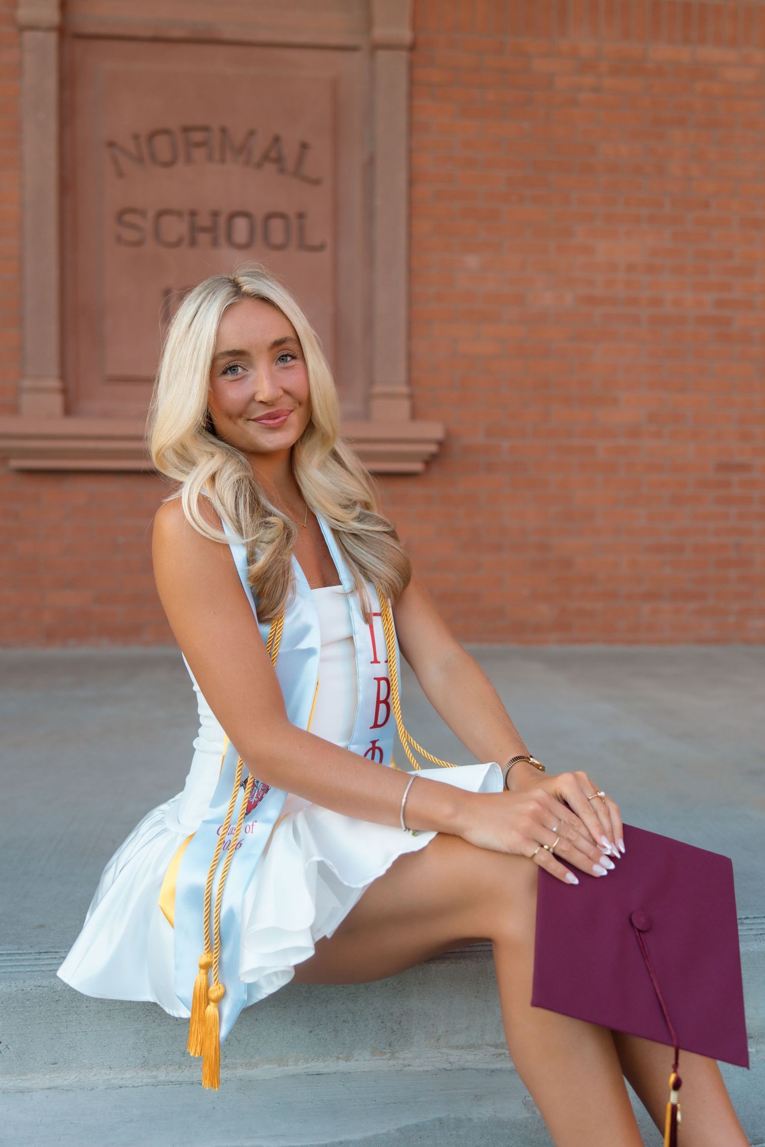 Blonde woman in white dress with graduation stoles sitting on steps outside Normal School building.