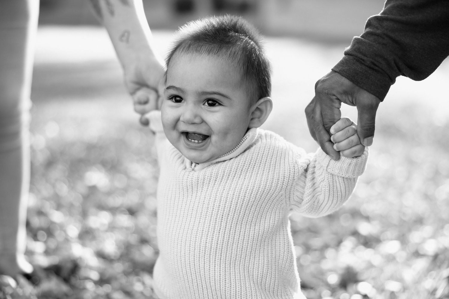 A joyful baby takes first steps holding parents hands outdoors in a black and white photo.