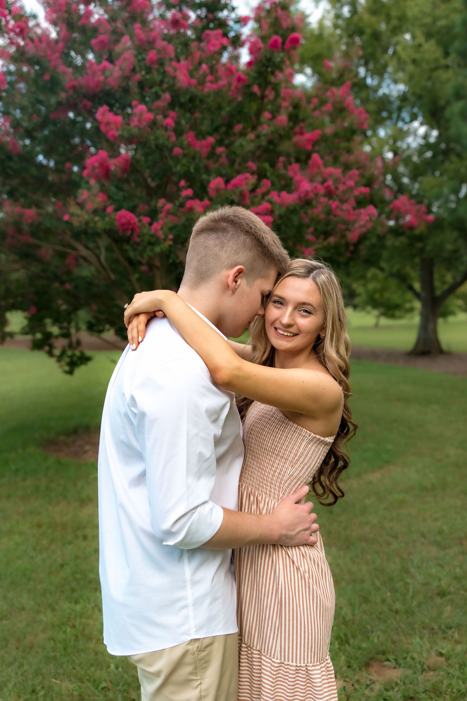Young couple embracing outdoors in a park, smiling woman in striped dress with long hair, pink flowering tree behind them.