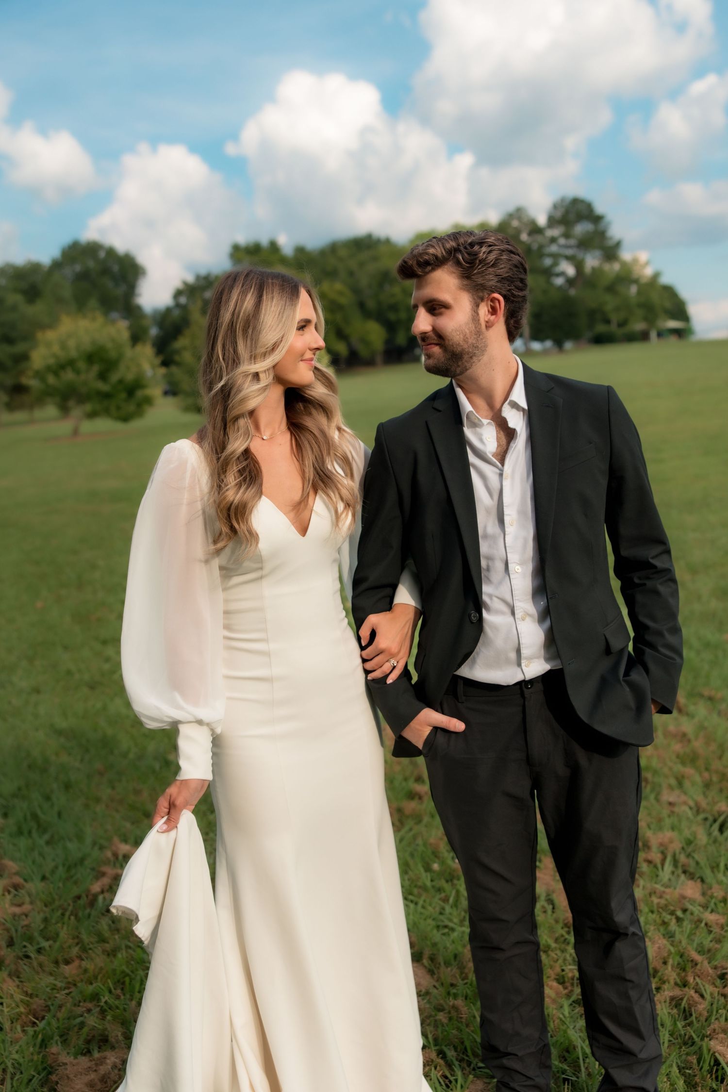 Bride and groom holding hands gazing at each other in a sunlit green field on their wedding day.