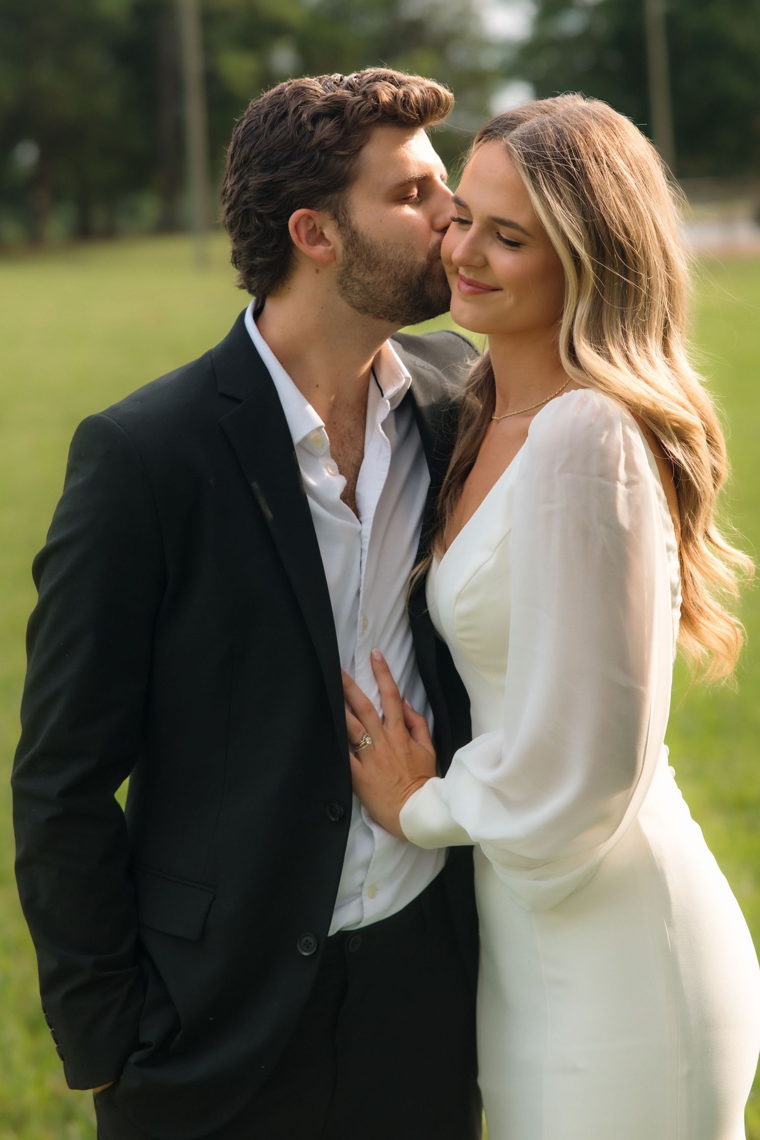 Groom in black suit kisses bride on cheek as she smiles softly, both standing in a sunlit green park.