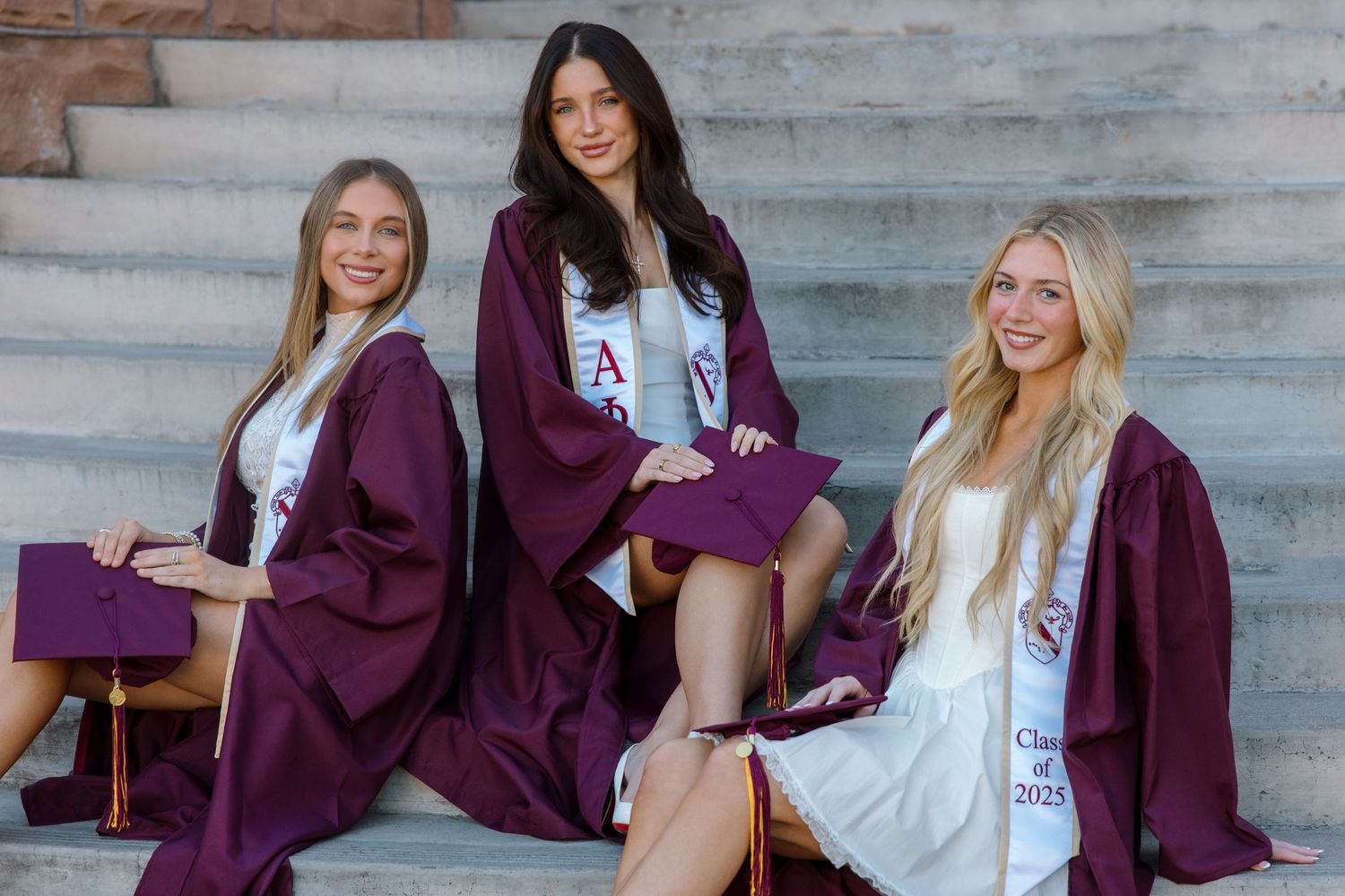 Three college graduates in maroon robes and Alpha Phi stoles sit on steps for graduation photos.