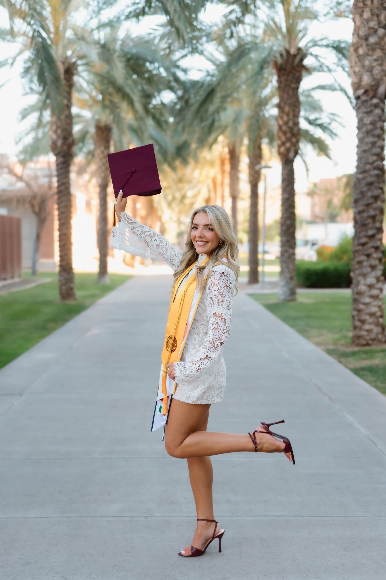 Joyful blonde graduate celebrates in lace dress and gold sash, raising maroon cap along palm tree-lined walkway.