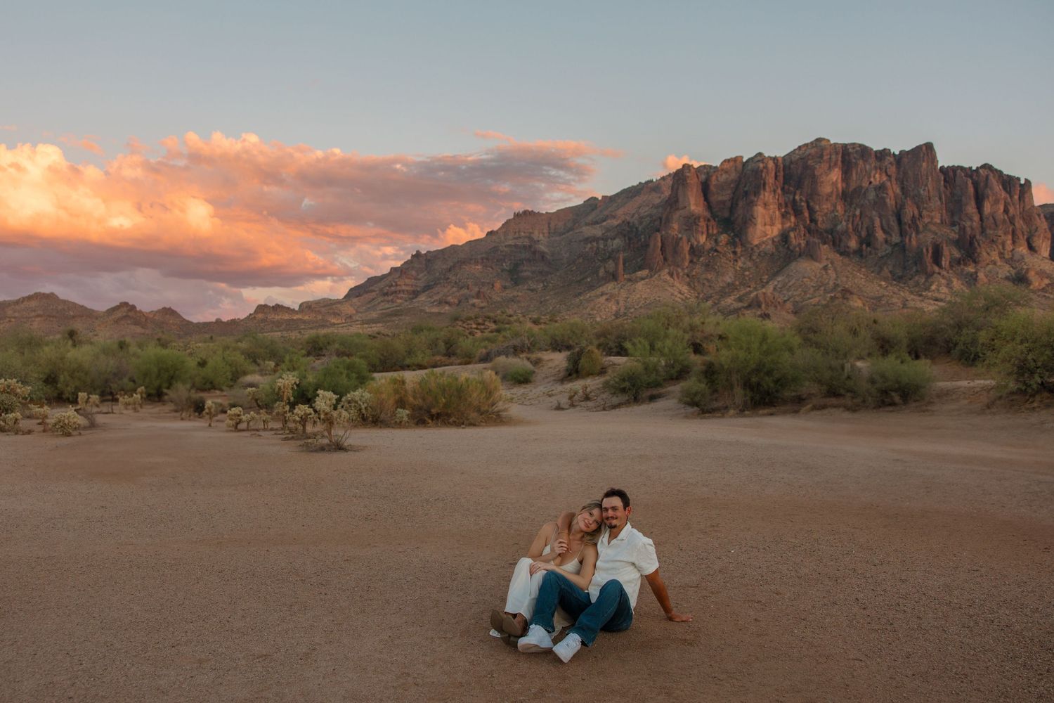Couple sitting together on desert ground at sunset with Superstition Mountains and colorful clouds in background.