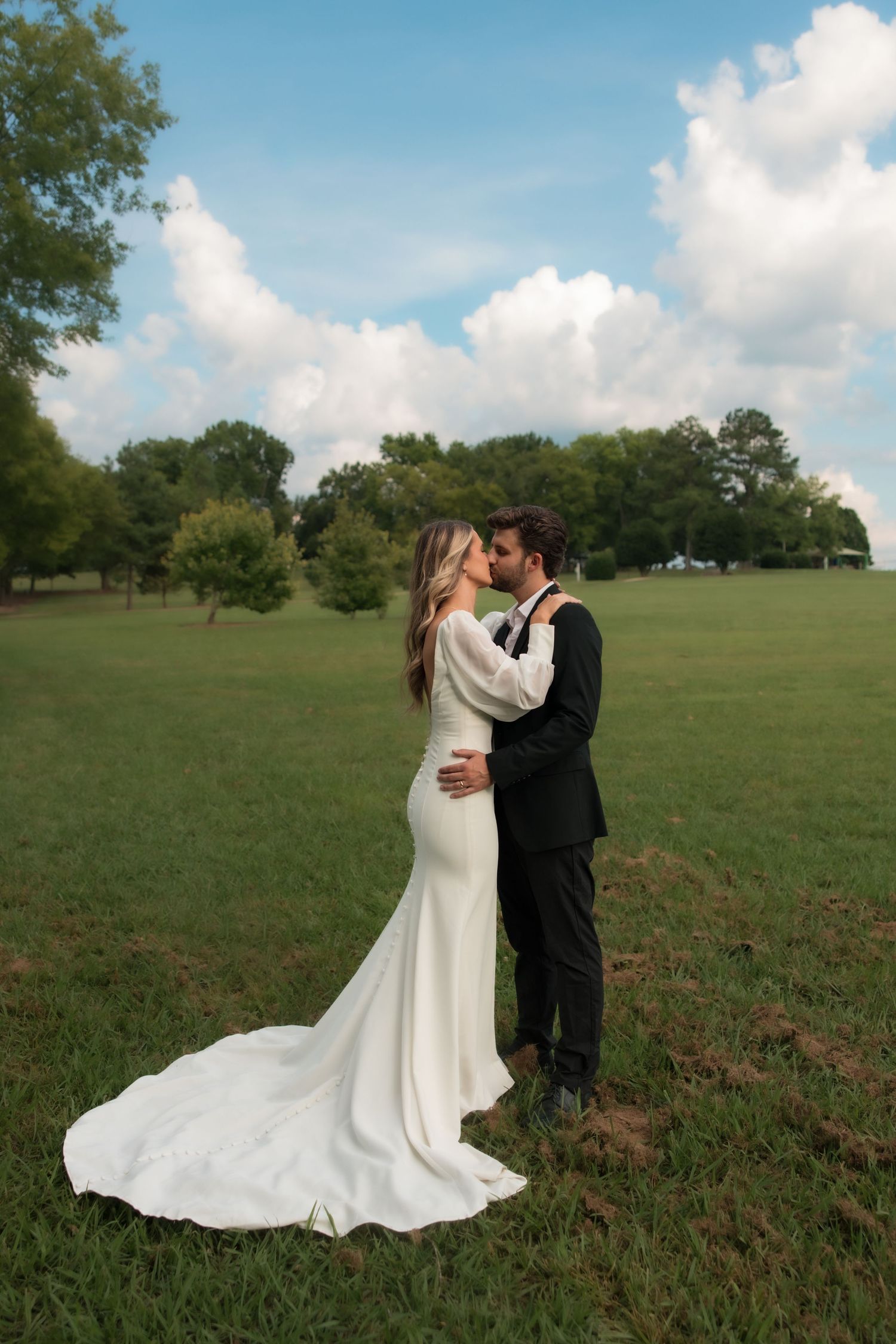 A bride and groom share a romantic kiss in an open green field under a dramatic cloudy sky.