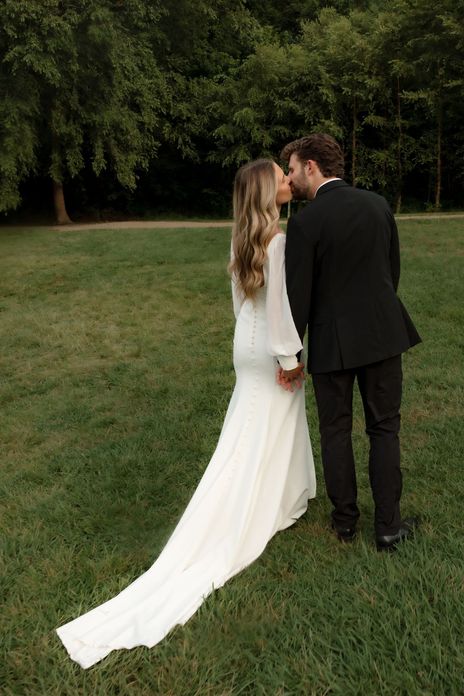 Bride and groom share a kiss while holding hands on green lawn with lush forest backdrop.