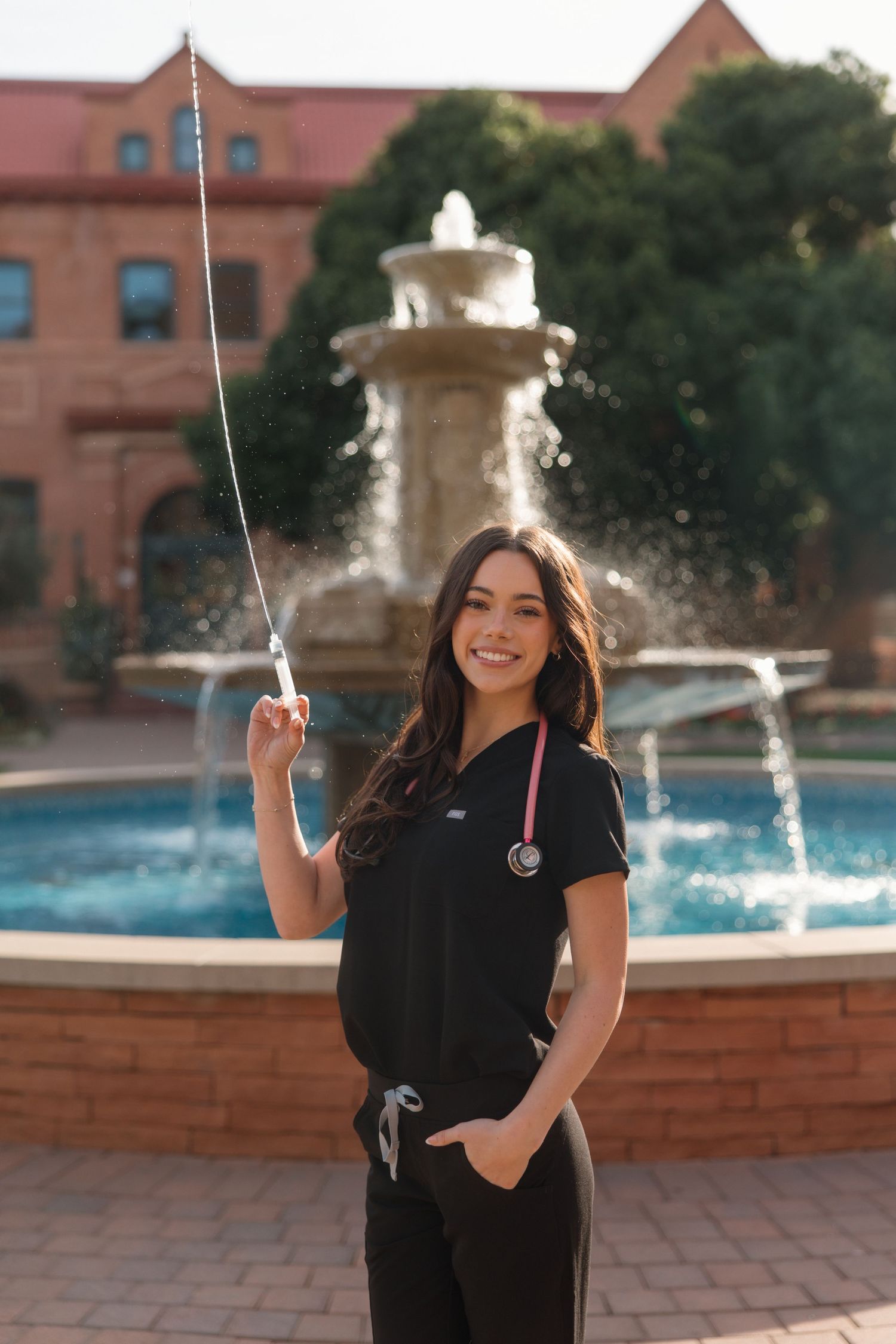 Smiling nursing graduate in black scrubs with pink stethoscope posing by fountain on college campus.
