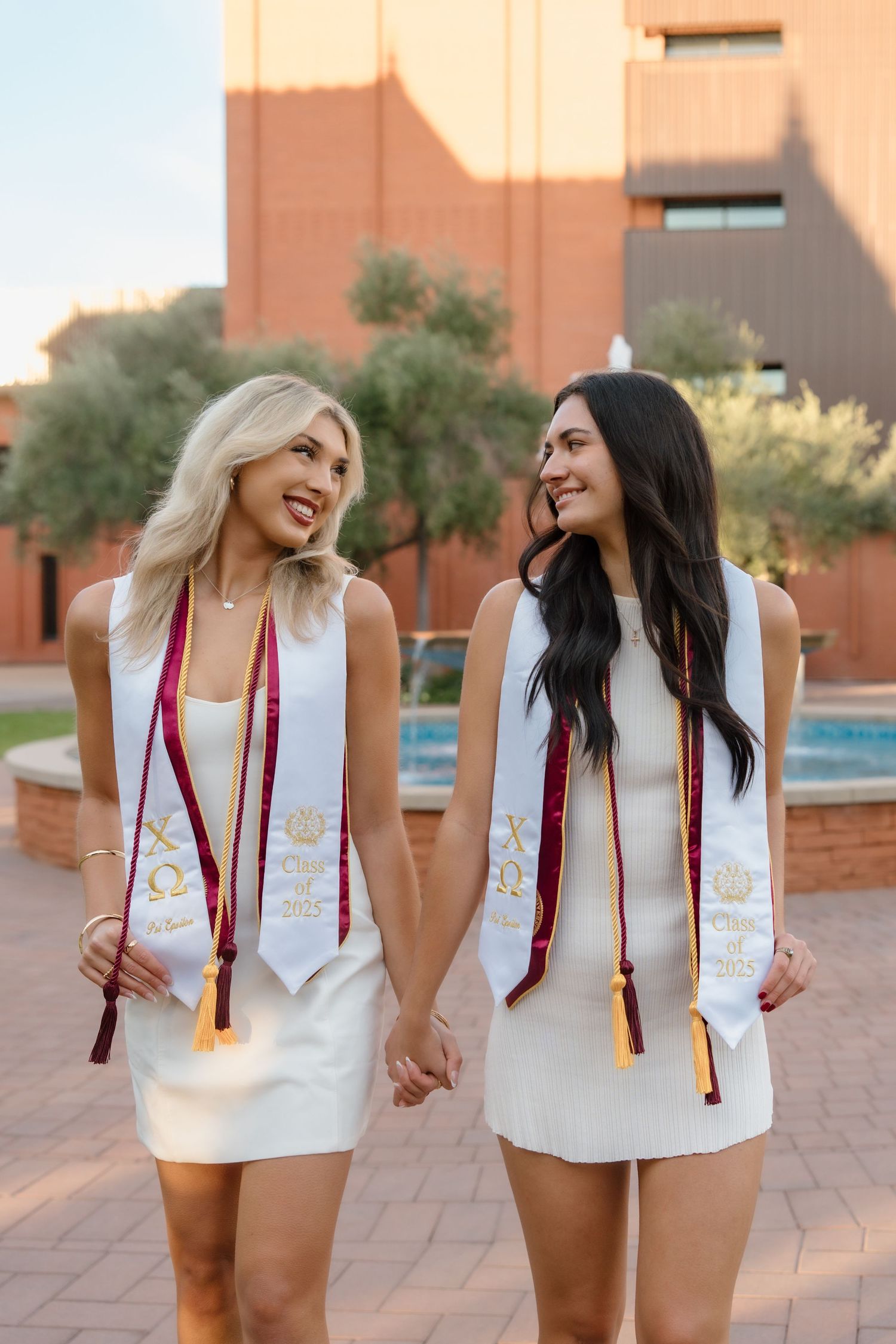 Two ASU graduates in white dresses and honor stoles holding hands and smiling while walking on campus.