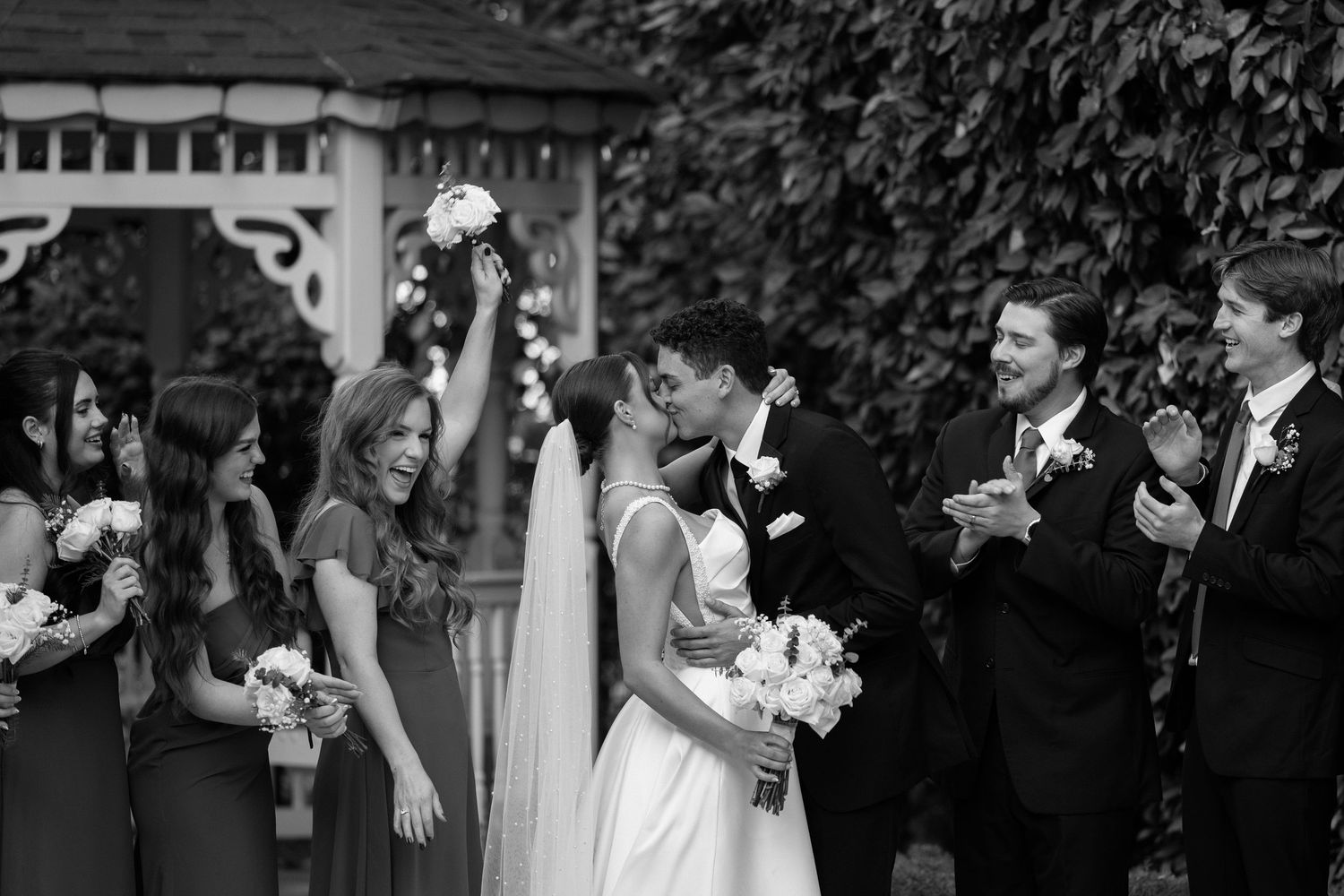 Black and white photo of bride and groom kissing as joyful wedding party celebrates at gazebo.
