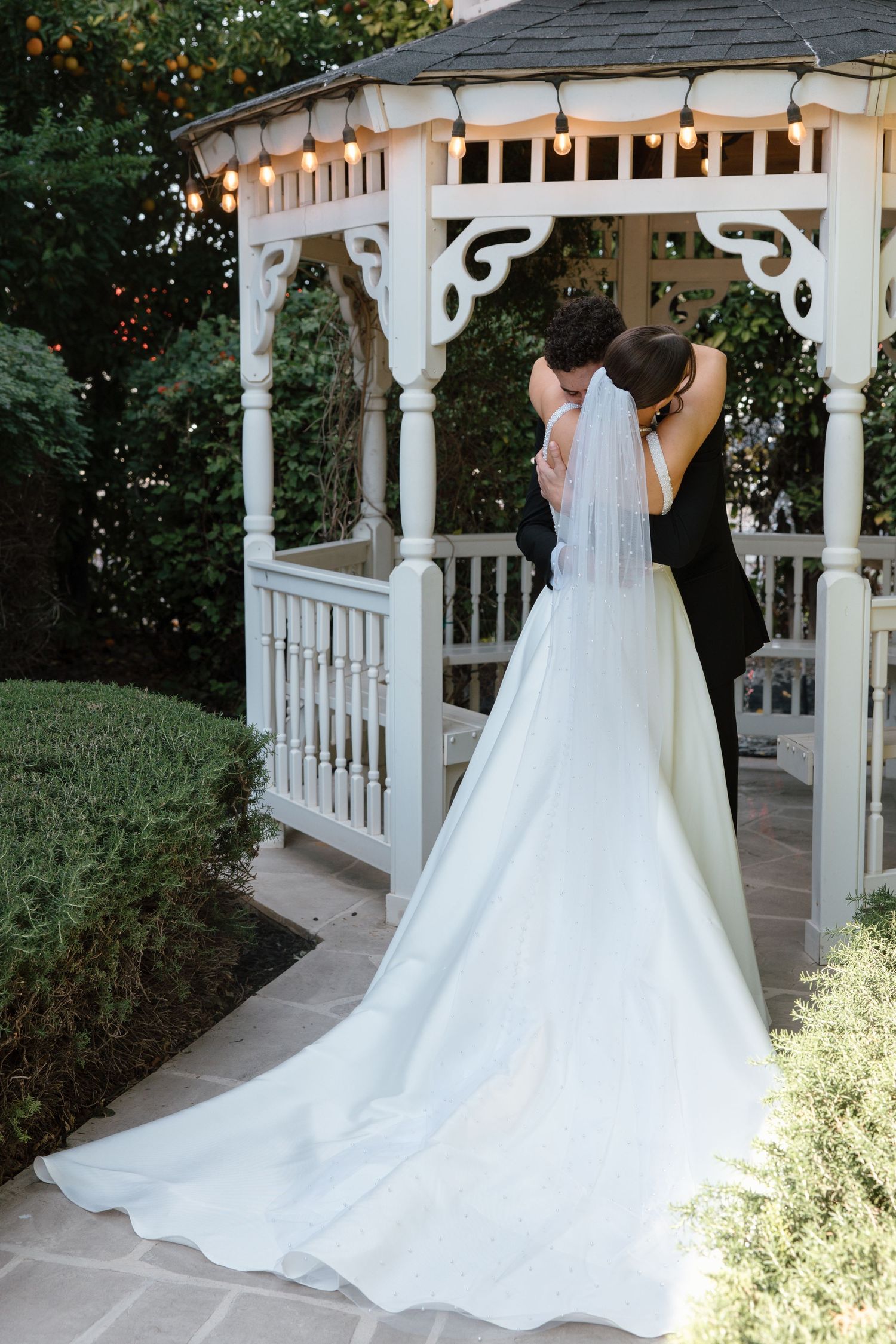 Bride in pearl-trimmed veil with long train embraces partner at elegant white garden gazebo.