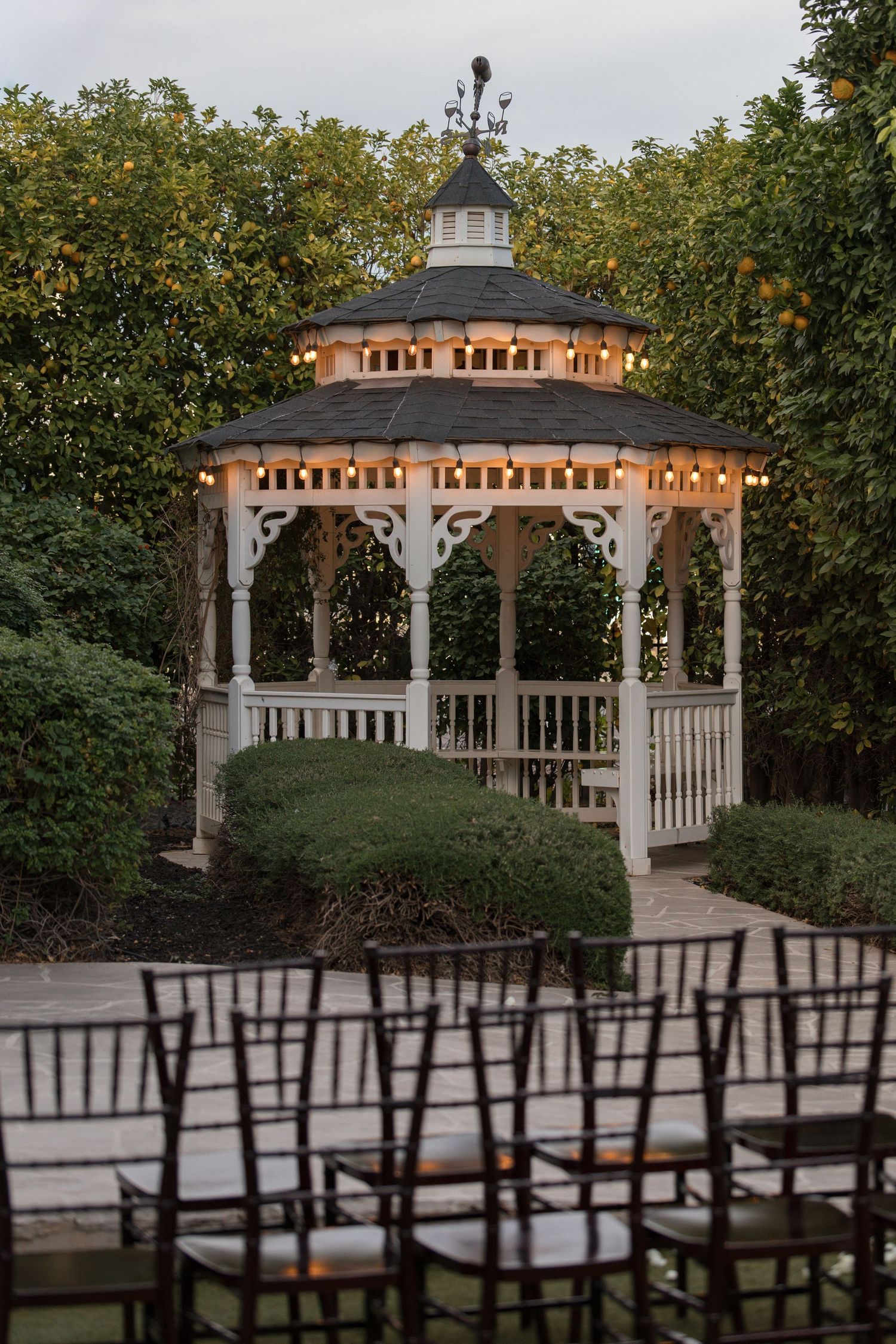 Charming white gazebo with warm string lights surrounded by lush greenery at dusk wedding venue.