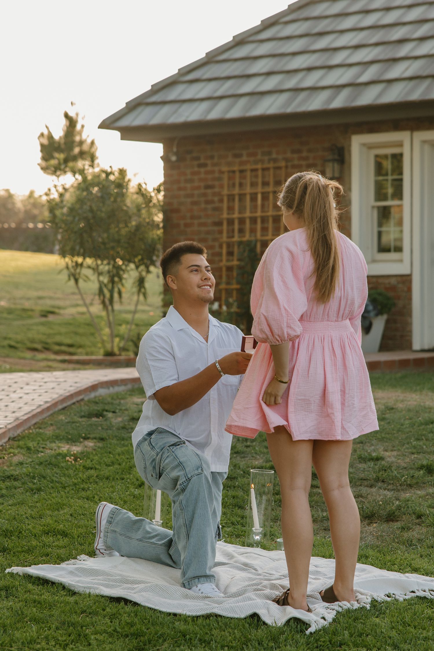Man on one knee proposing to woman in pink dress on blanket in garden at sunset near brick house.