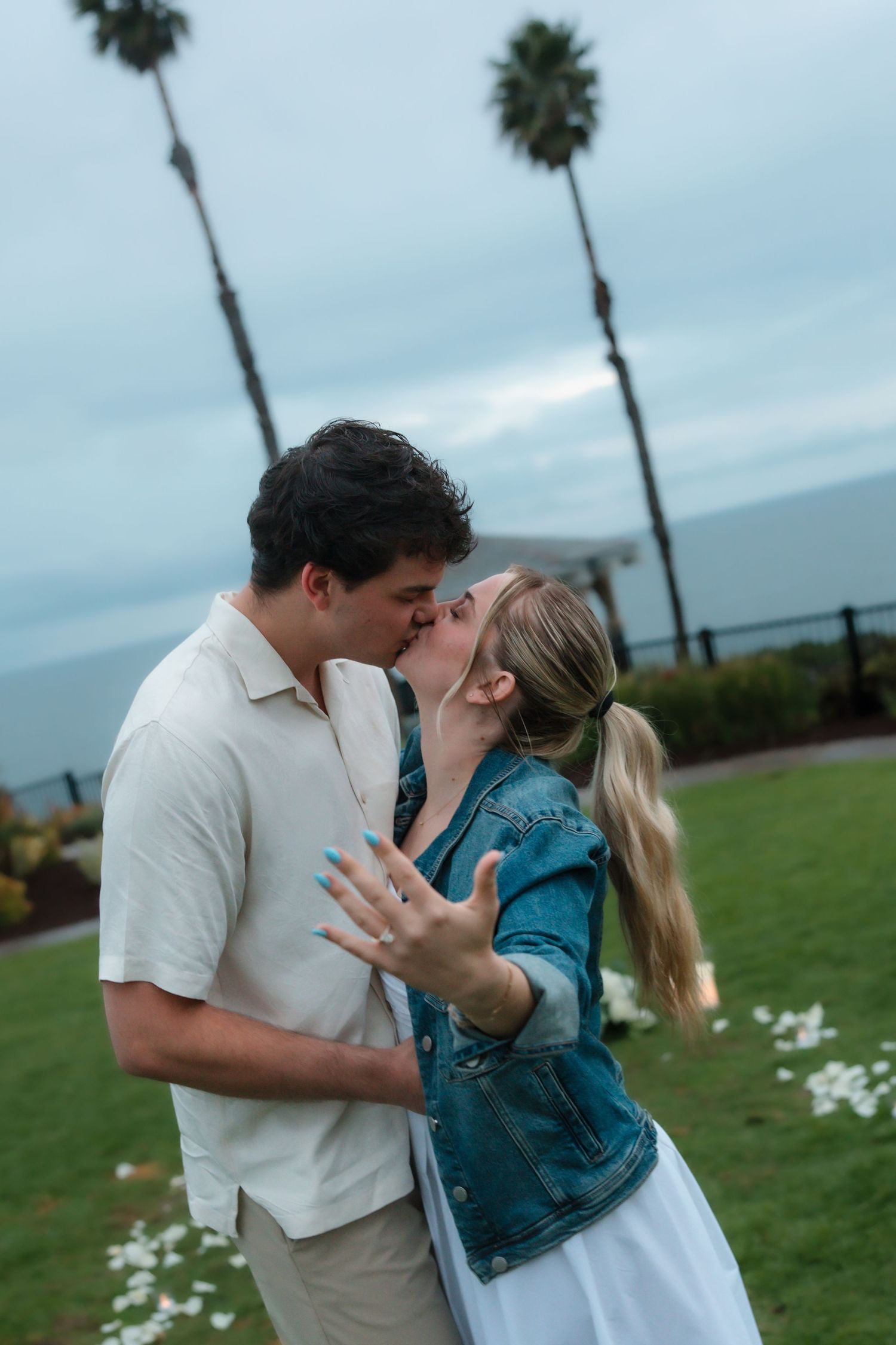 Newly engaged couple kissing outdoors, woman showing diamond ring with palm trees behind.