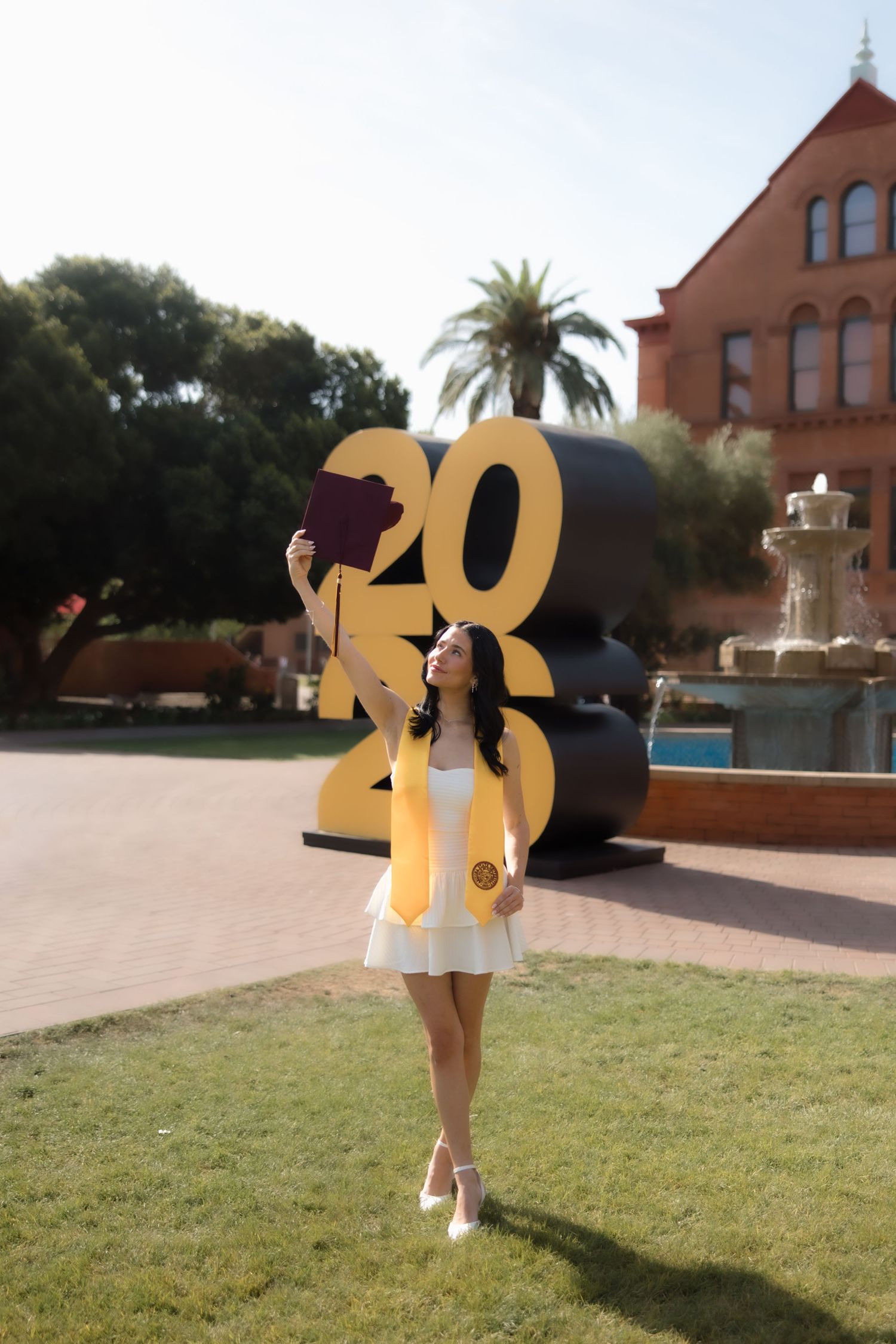 A young woman in a white dress and gold graduation stole holds her cap by a 2020 sign on a sunny campus.
