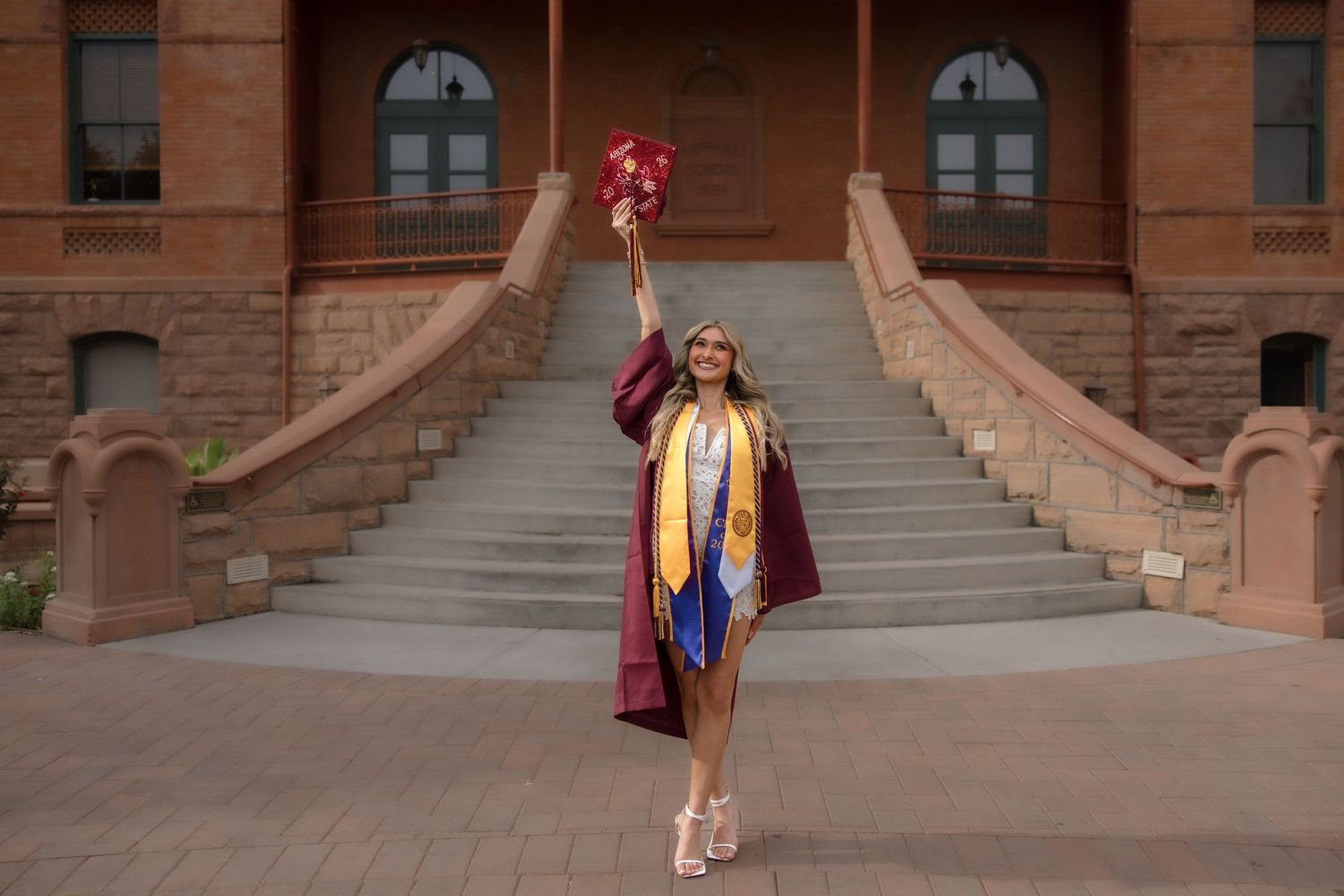 Graduate in maroon cap and gown raises tassel bouquet triumphantly in front of historic brick university building steps.