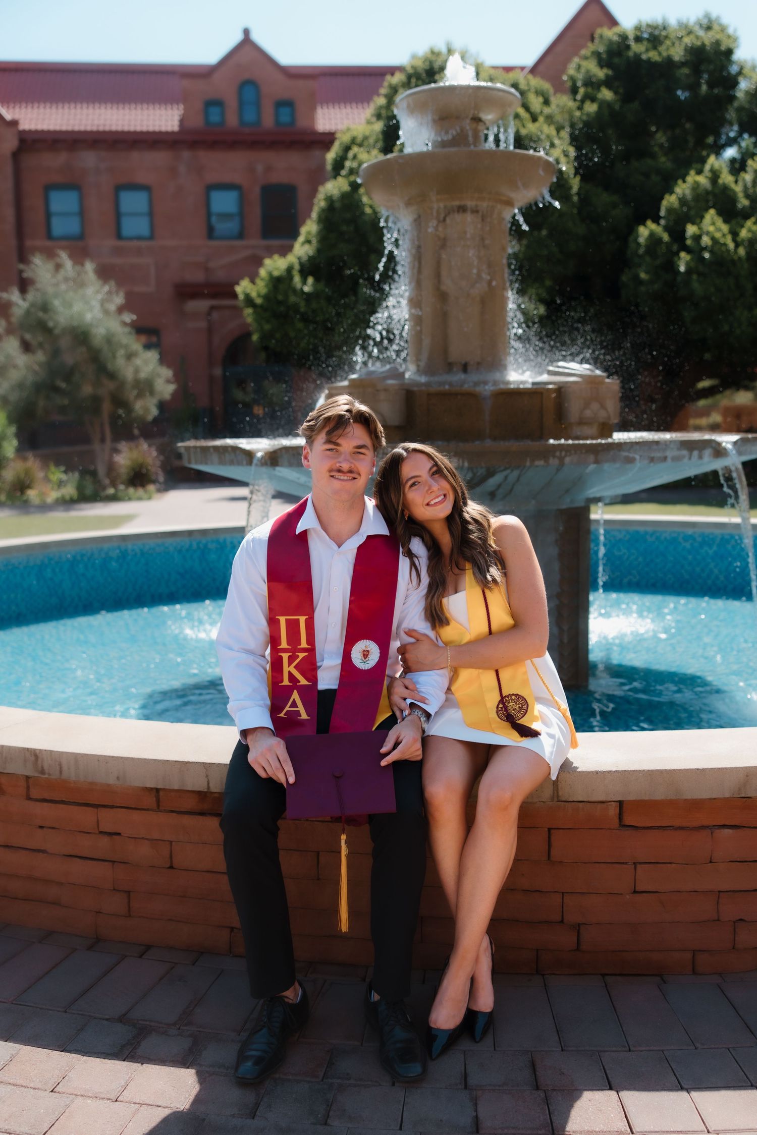 Two college graduates in regalia sit smiling by a fountain on campus, one wearing a Pi Kappa Alpha stole.