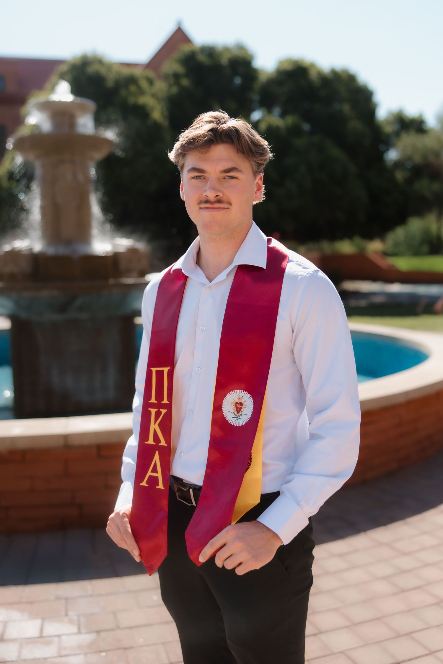 Male graduate wearing Pi Kappa Alpha fraternity stole posing by campus fountain at ASU.