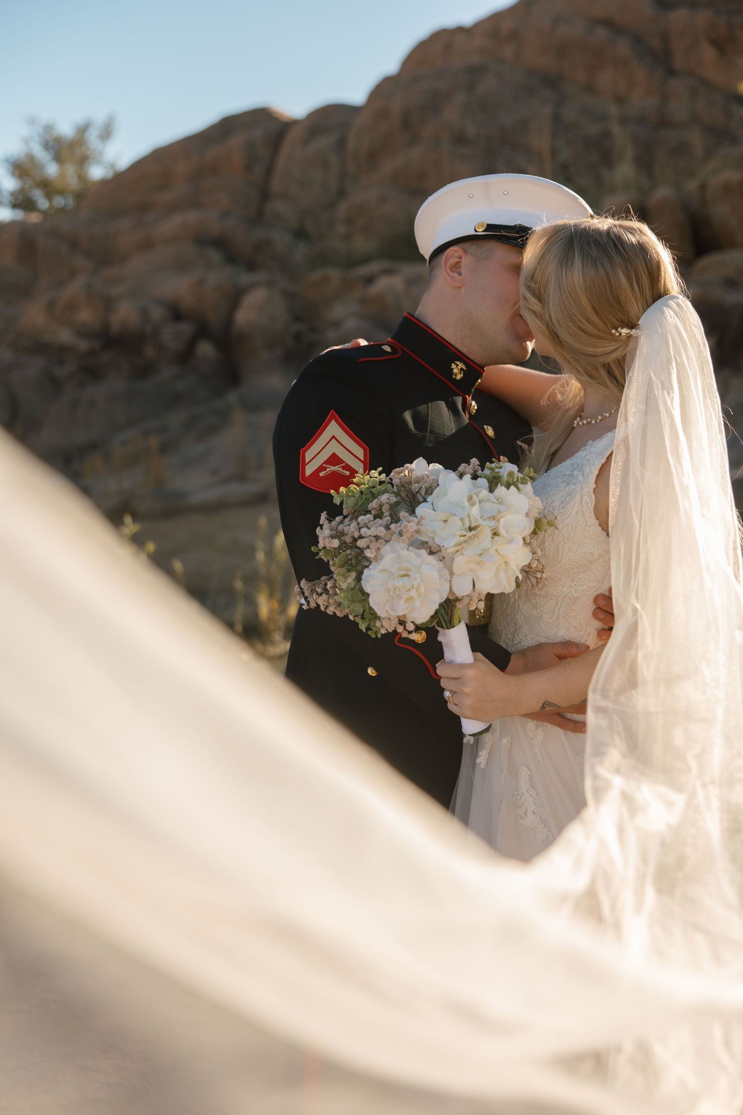 Marine groom and bride share a kiss with billowing veil in foreground and rocky desert backdrop.