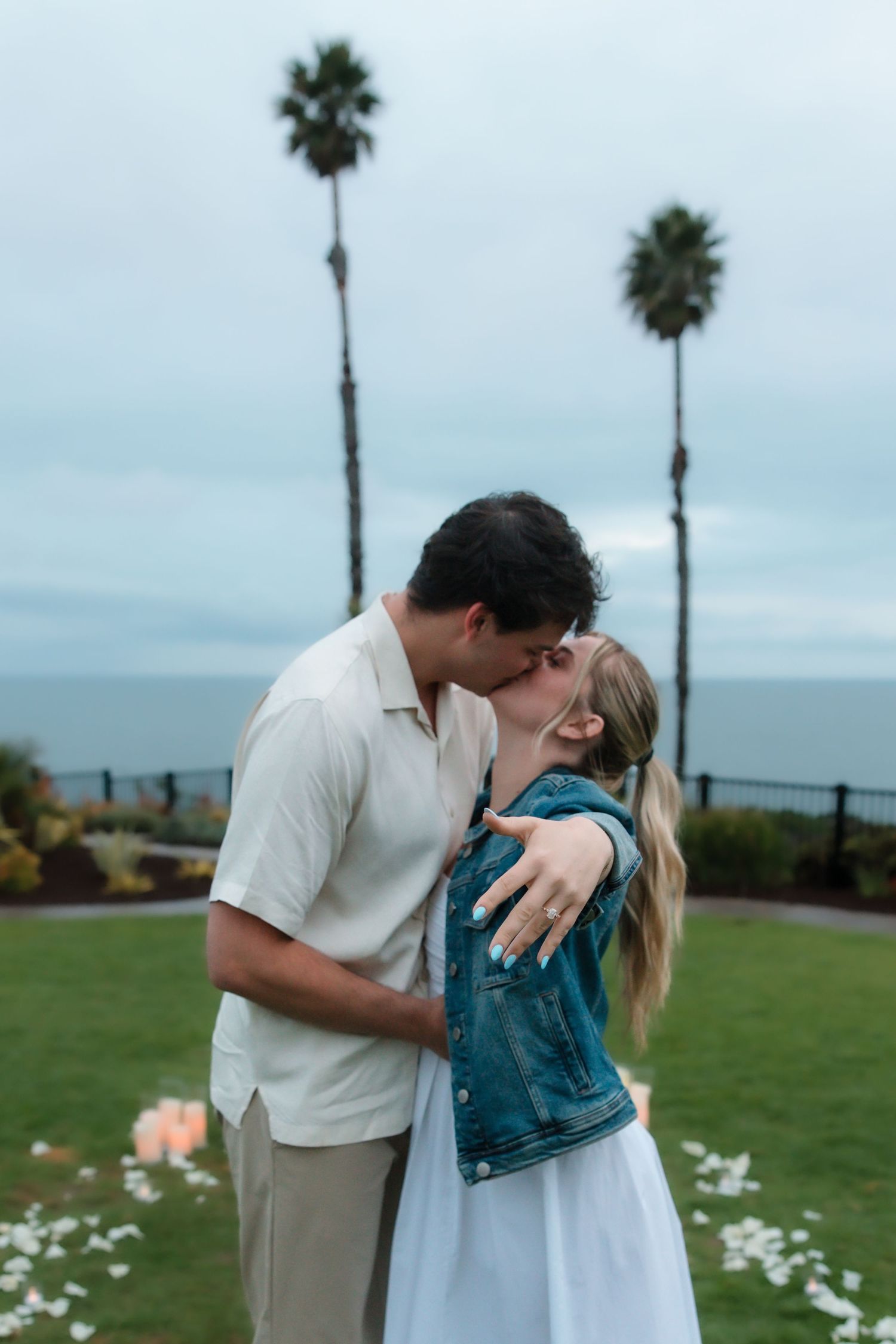 Newly engaged couple kissing outdoors showing engagement ring with palm trees and ocean backdrop.
