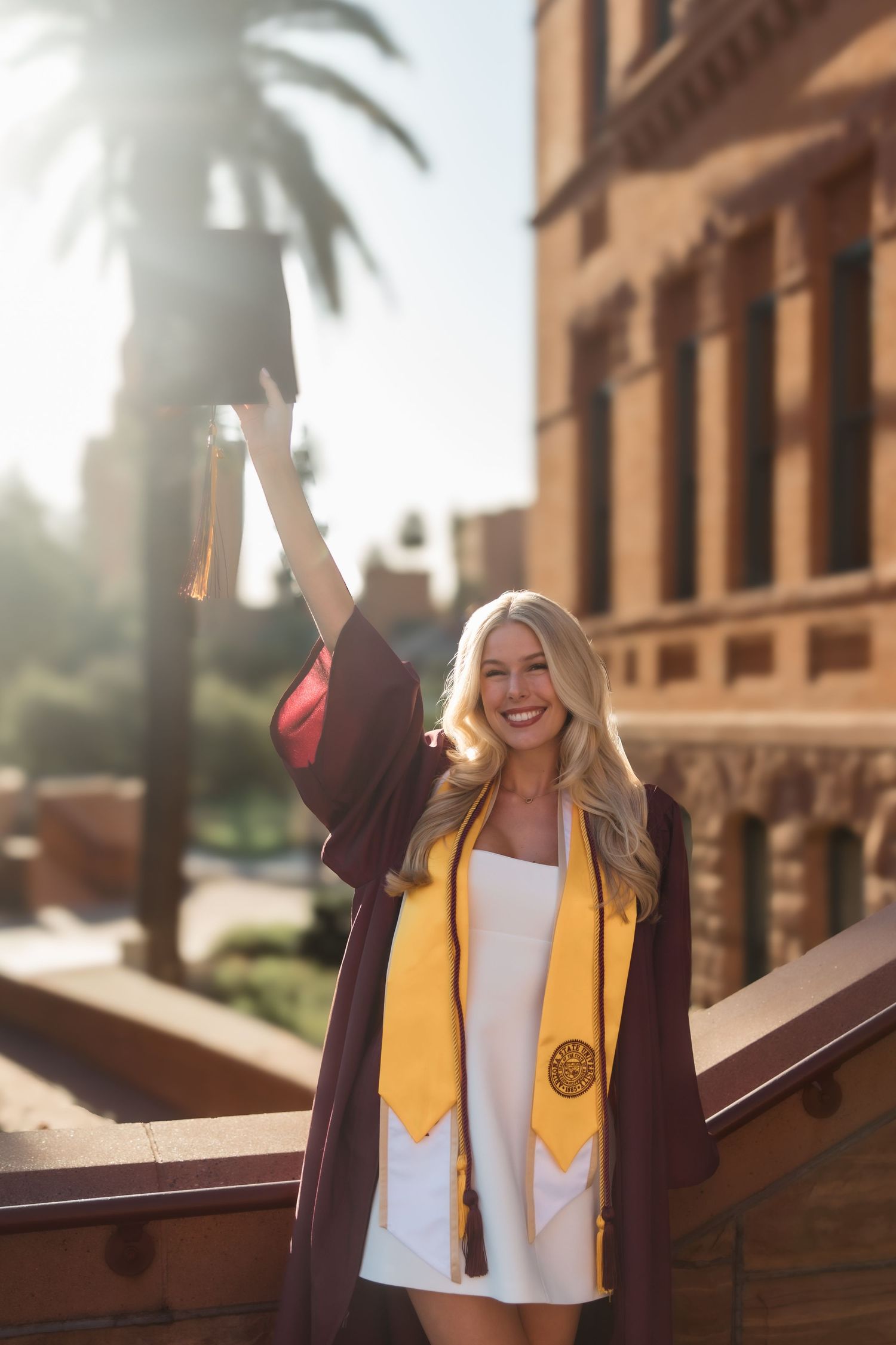 Smiling female graduate raises cap wearing maroon gown with gold ASU stole on sunlit campus balcony with palm tree.