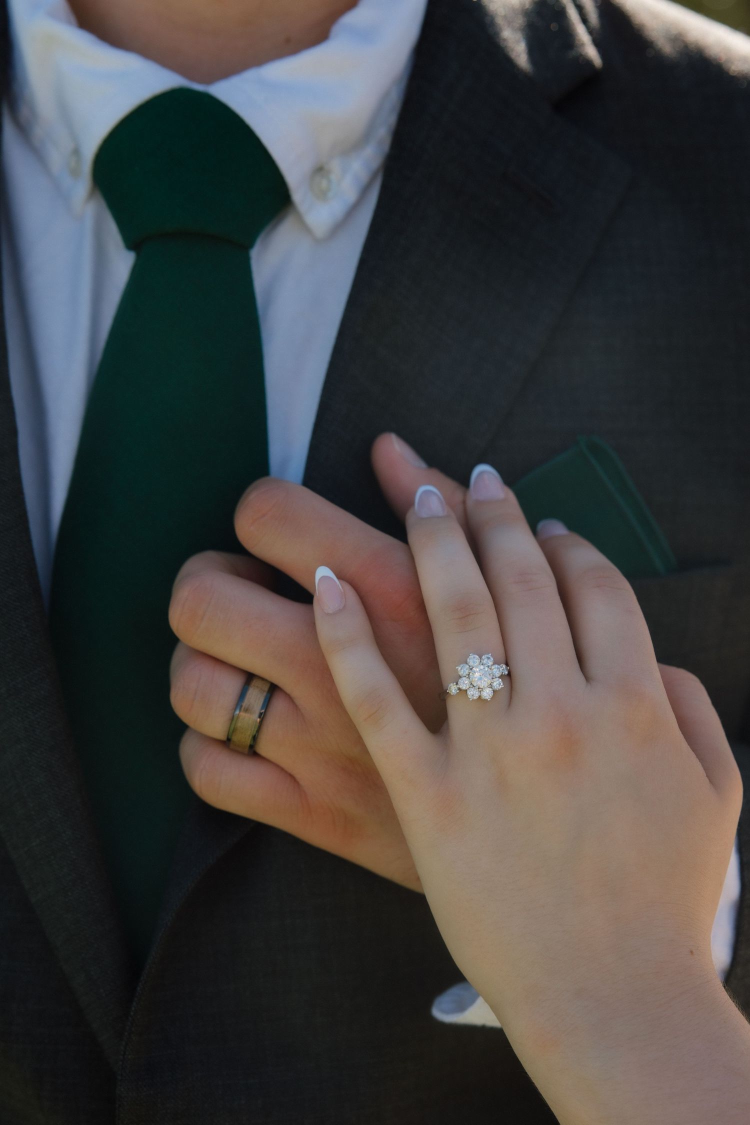 Close-up of a bride's hand displaying a floral diamond engagement ring resting on a groom's dark suit with green tie.