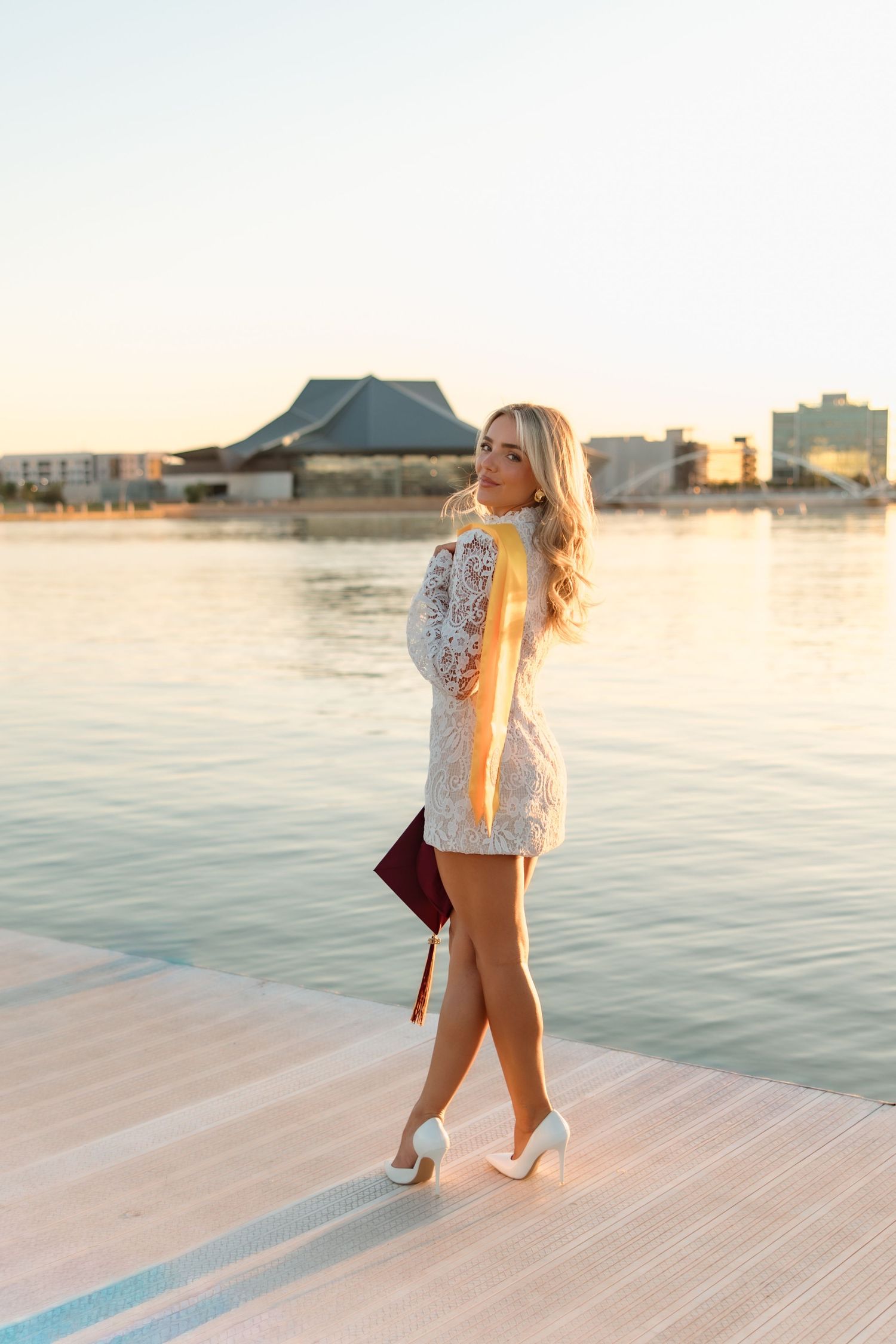 Blonde woman in white lace mini dress and heels walks along waterfront dock at golden sunset.