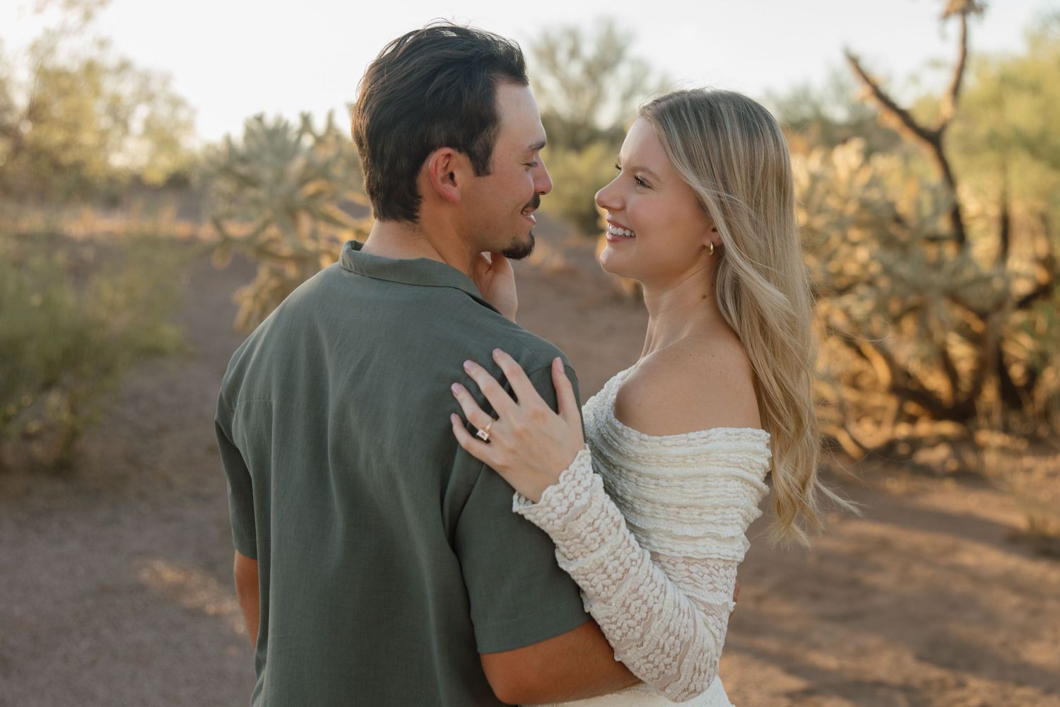 Couple embracing and smiling in a desert landscape, woman in white lace dress, man in olive green shirt.