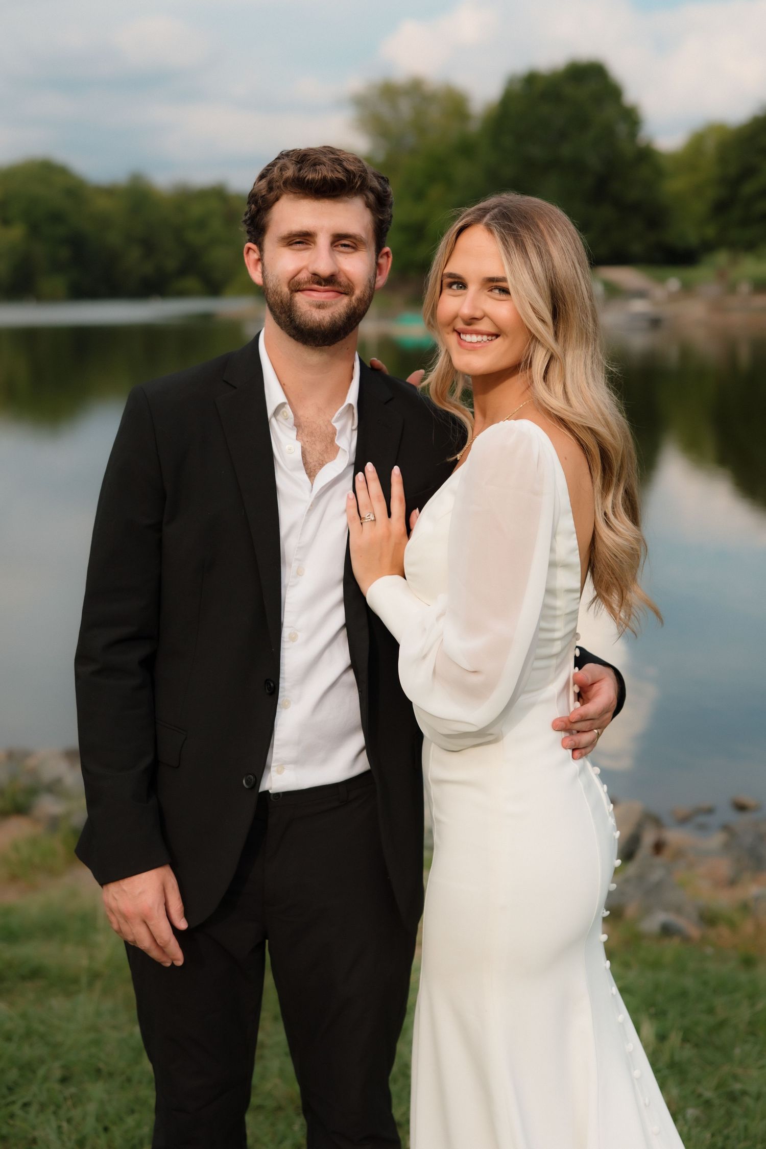Smiling couple poses near a lake, bride in white long-sleeve gown showing ring, groom in black suit.