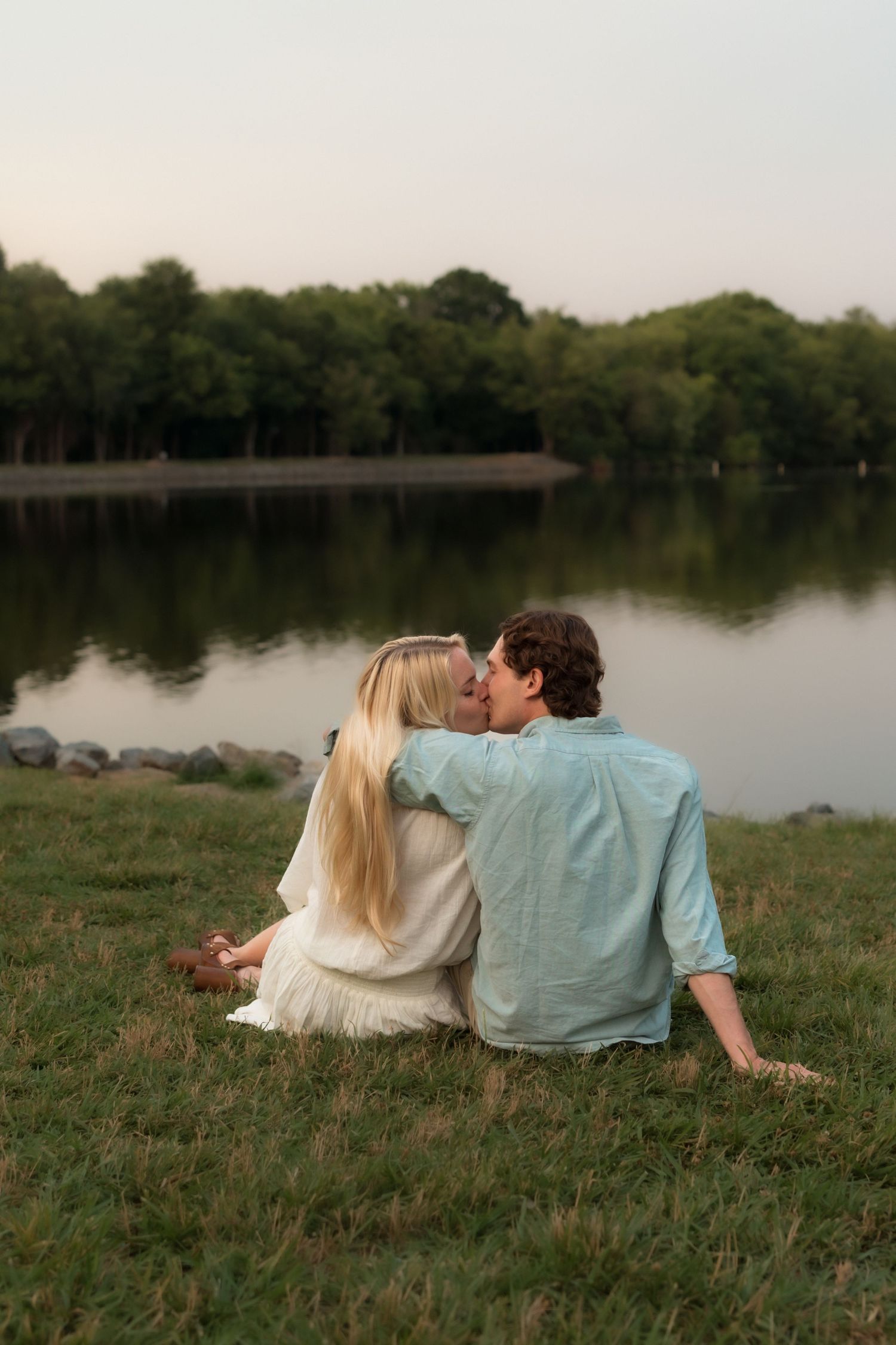 Couple sharing a romantic kiss while sitting on grass beside a peaceful lake at dusk.