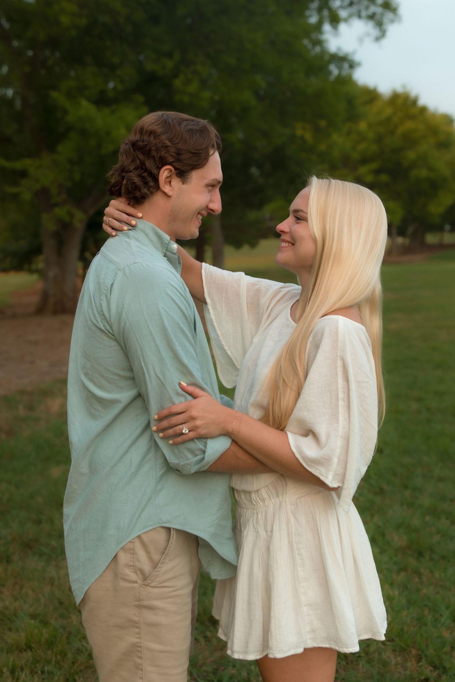 Engaged couple embracing and smiling at each other in a green outdoor setting.
