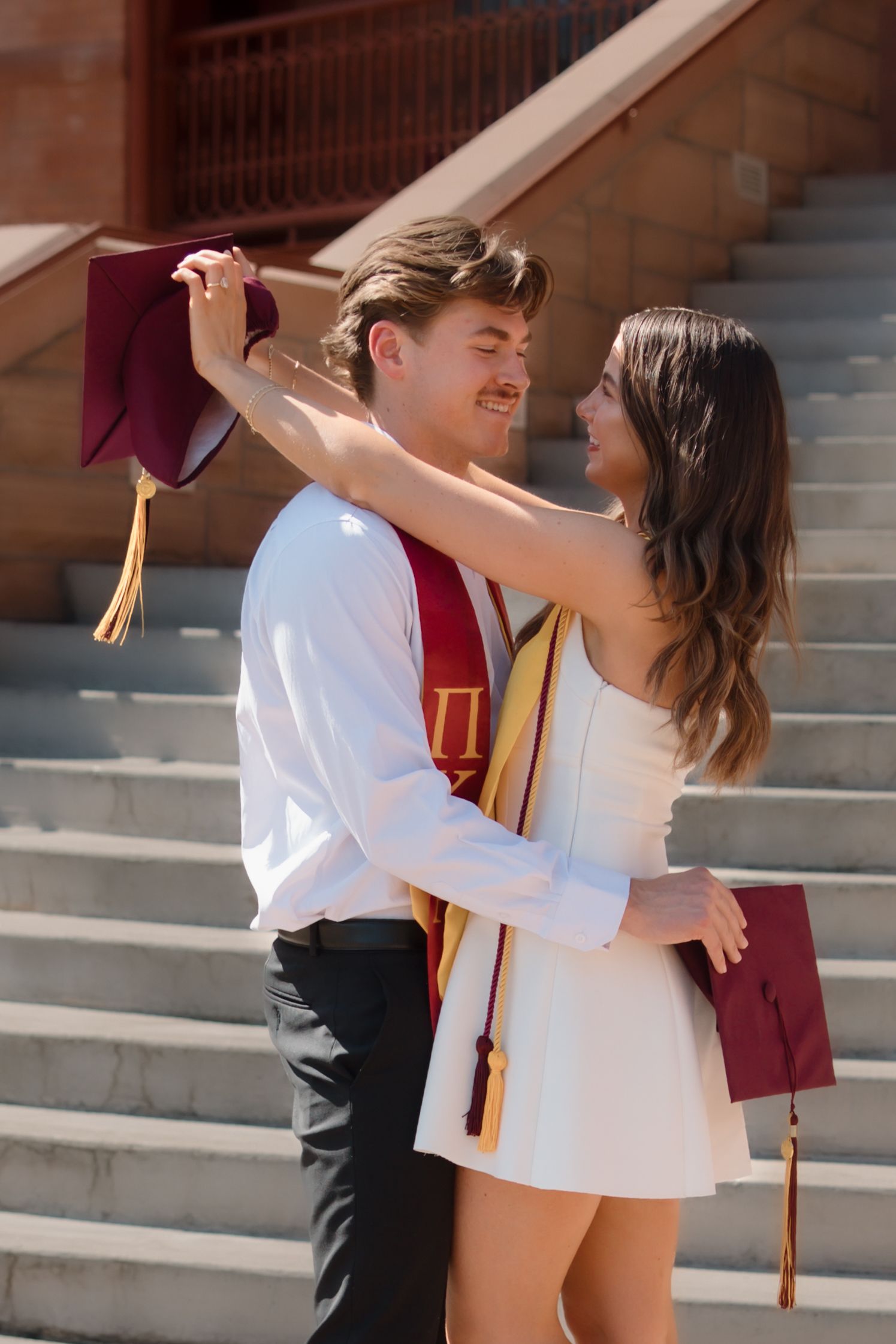 Joyful graduating couple embracing on campus steps wearing Pi Kappa Alpha and gold graduation stoles.