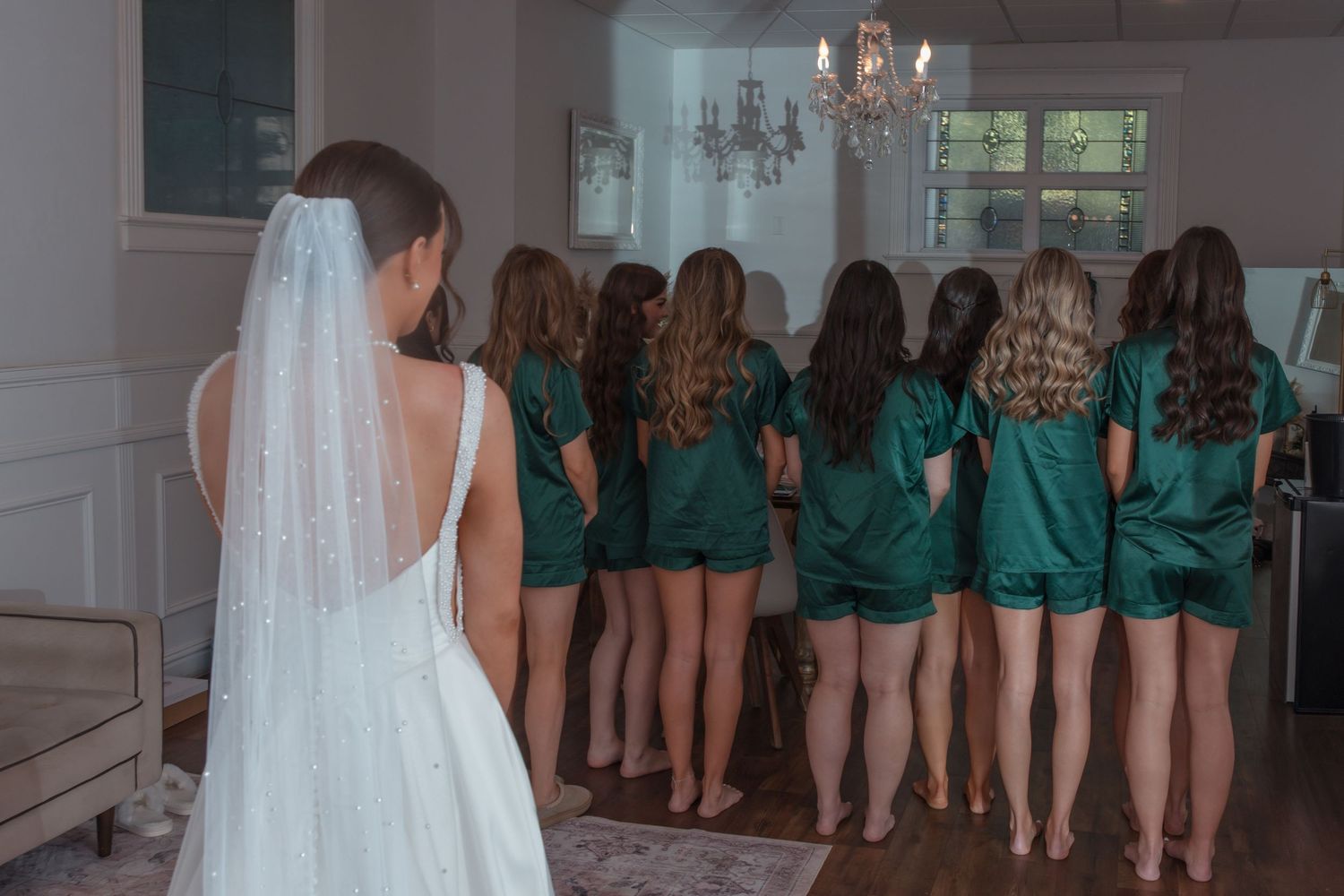 Bride in white gown and veil approaching bridesmaids in emerald green satin pajamas for first look.