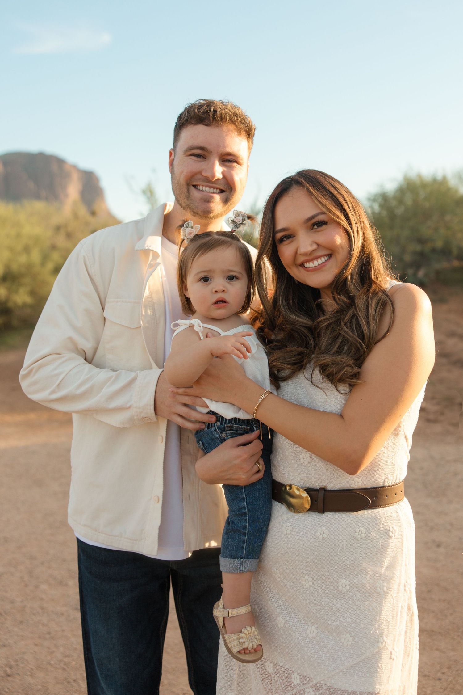 Smiling young family with baby girl posing outdoors with desert mountains in the background.