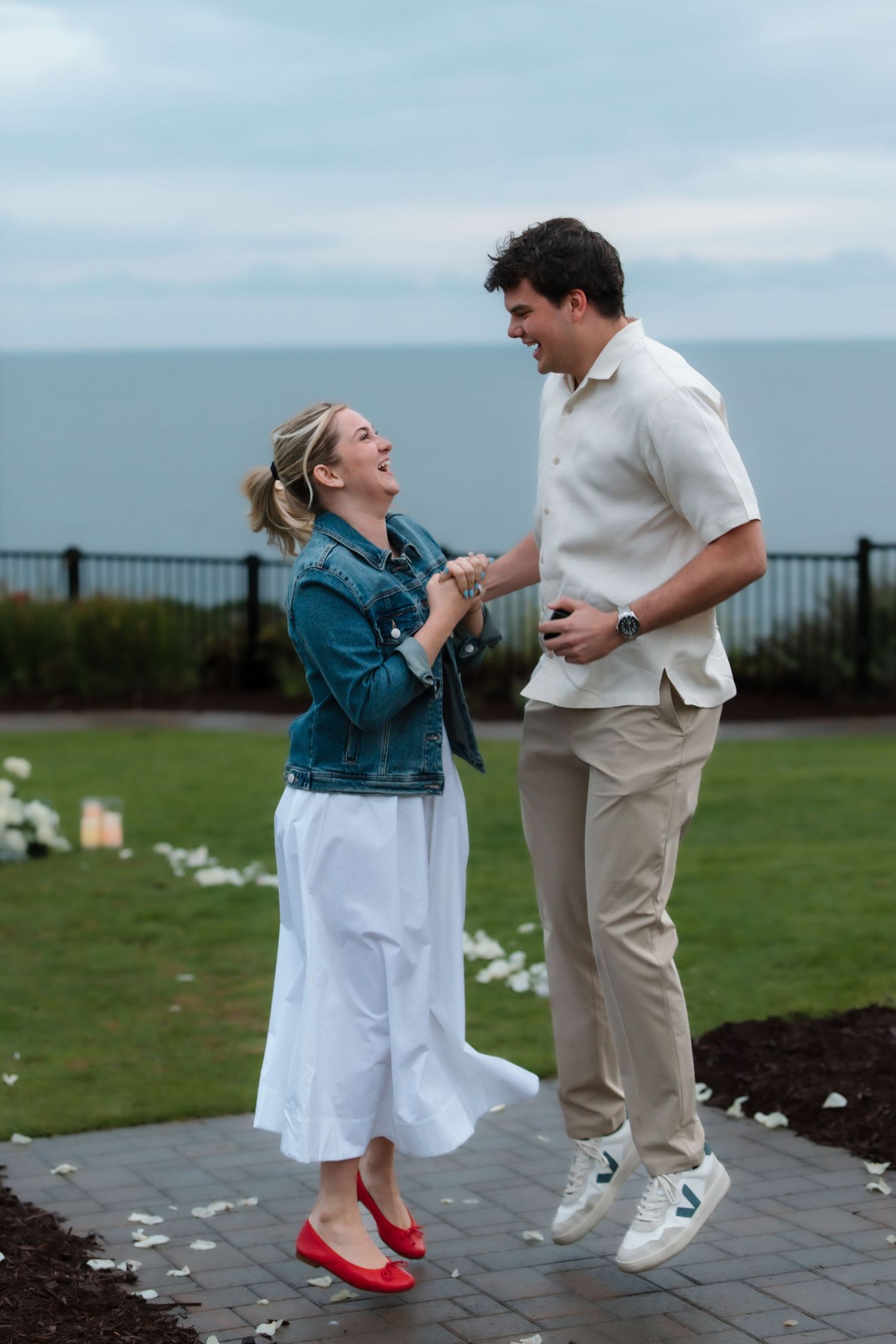 Joyful newly engaged couple laughing and jumping together on coastal garden pathway.