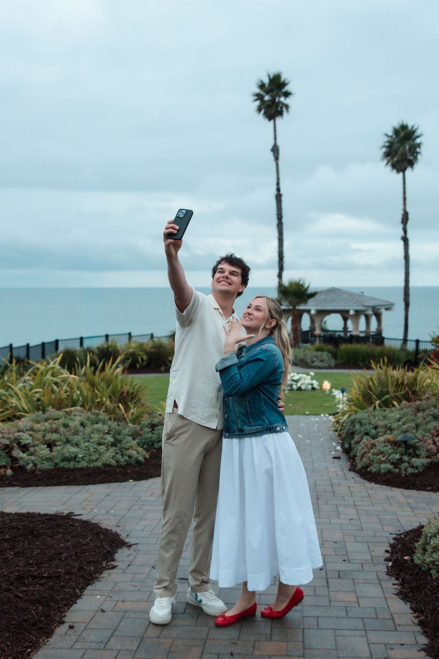 Newly engaged couple taking selfie in scenic garden with palm trees and ocean backdrop.