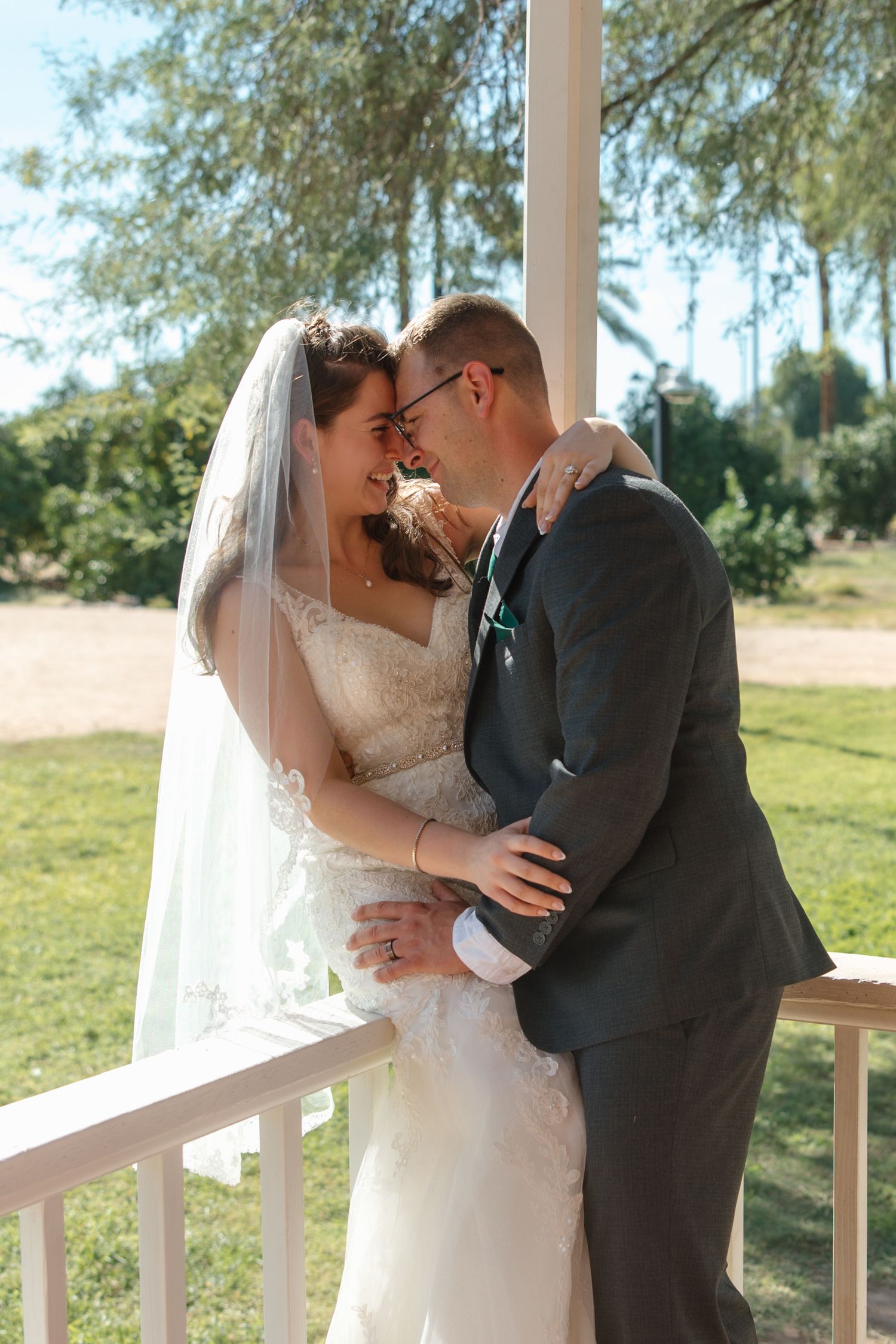 Bride and groom embrace tenderly on a white porch gazebo surrounded by lush greenery on their wedding day.