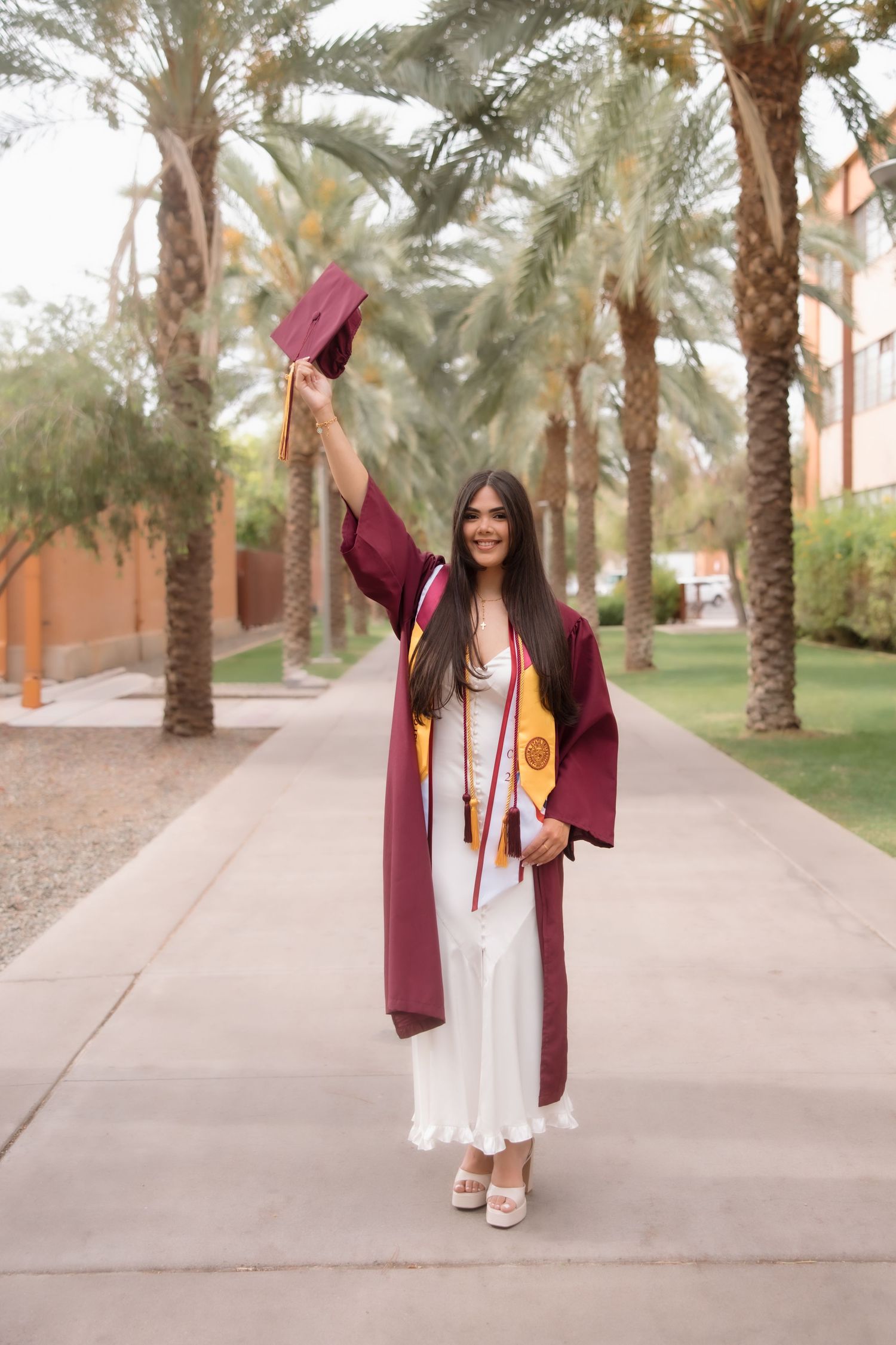 Young woman in maroon graduation gown and cap raised high, celebrating outdoors among palm trees on a sunny campus.