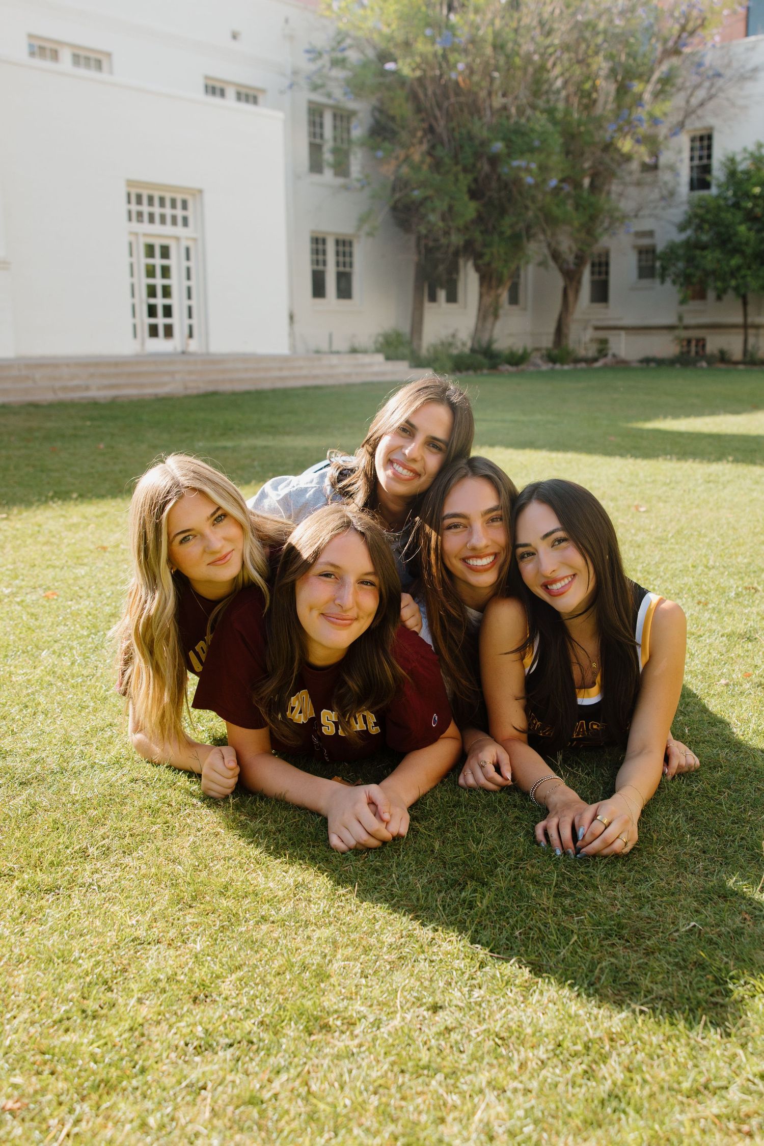 Five young women pose together smiling on green grass lawn in front of university building.
