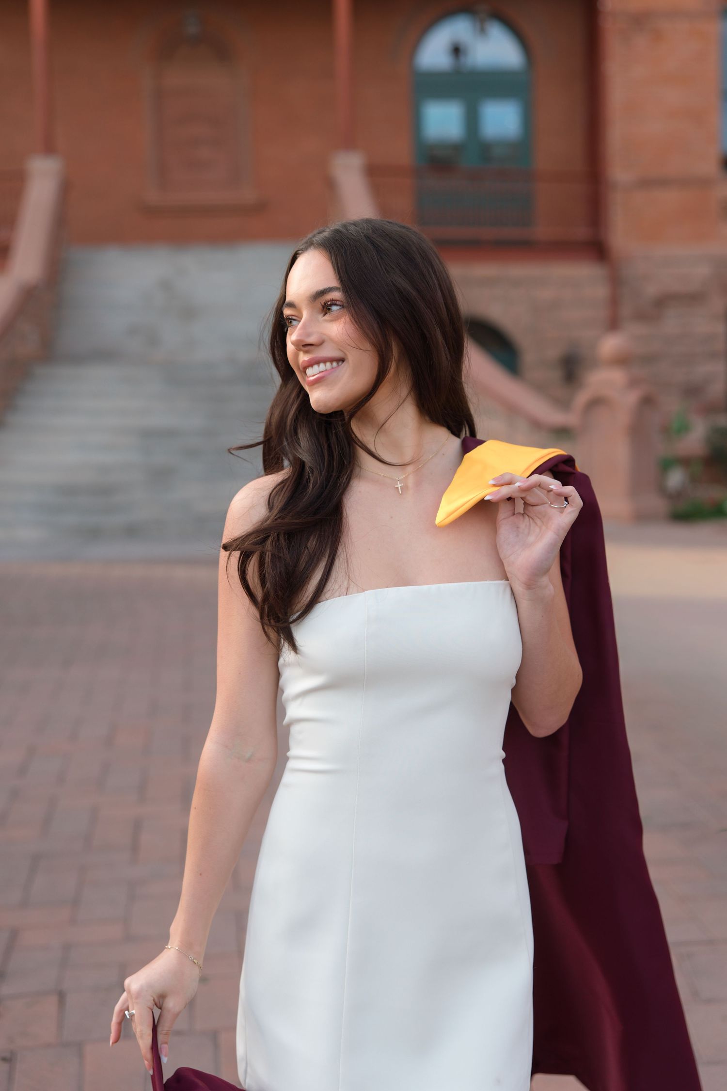 Female graduate in white strapless dress carrying gold and maroon graduation regalia on campus steps.