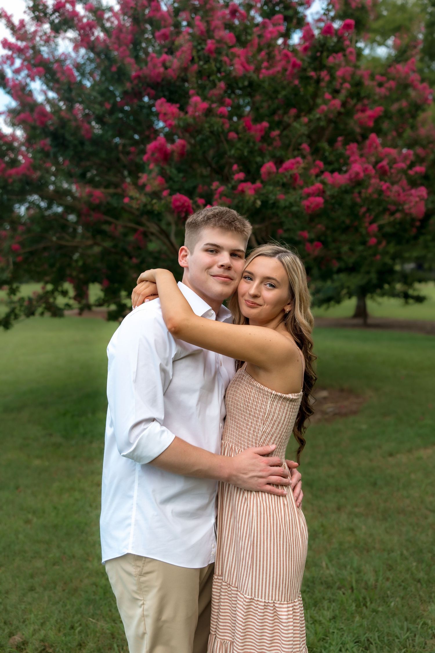 Young couple embracing outdoors in front of a blooming pink crepe myrtle tree on a green lawn.