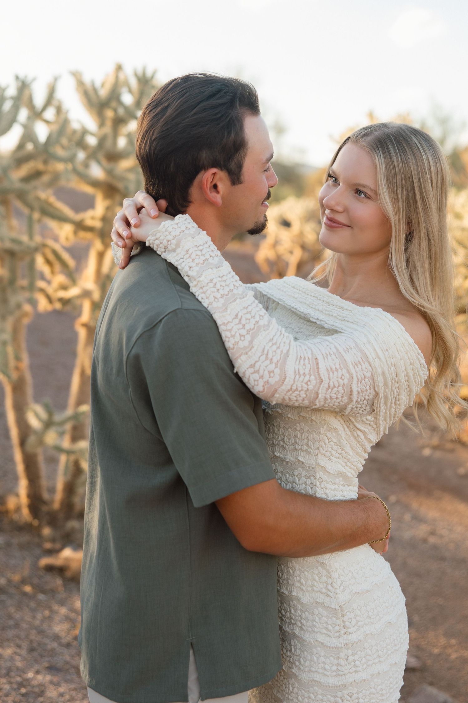 Couple embracing in desert landscape with cacti, woman in white knit dress smiling, man in olive shirt, golden hour light.