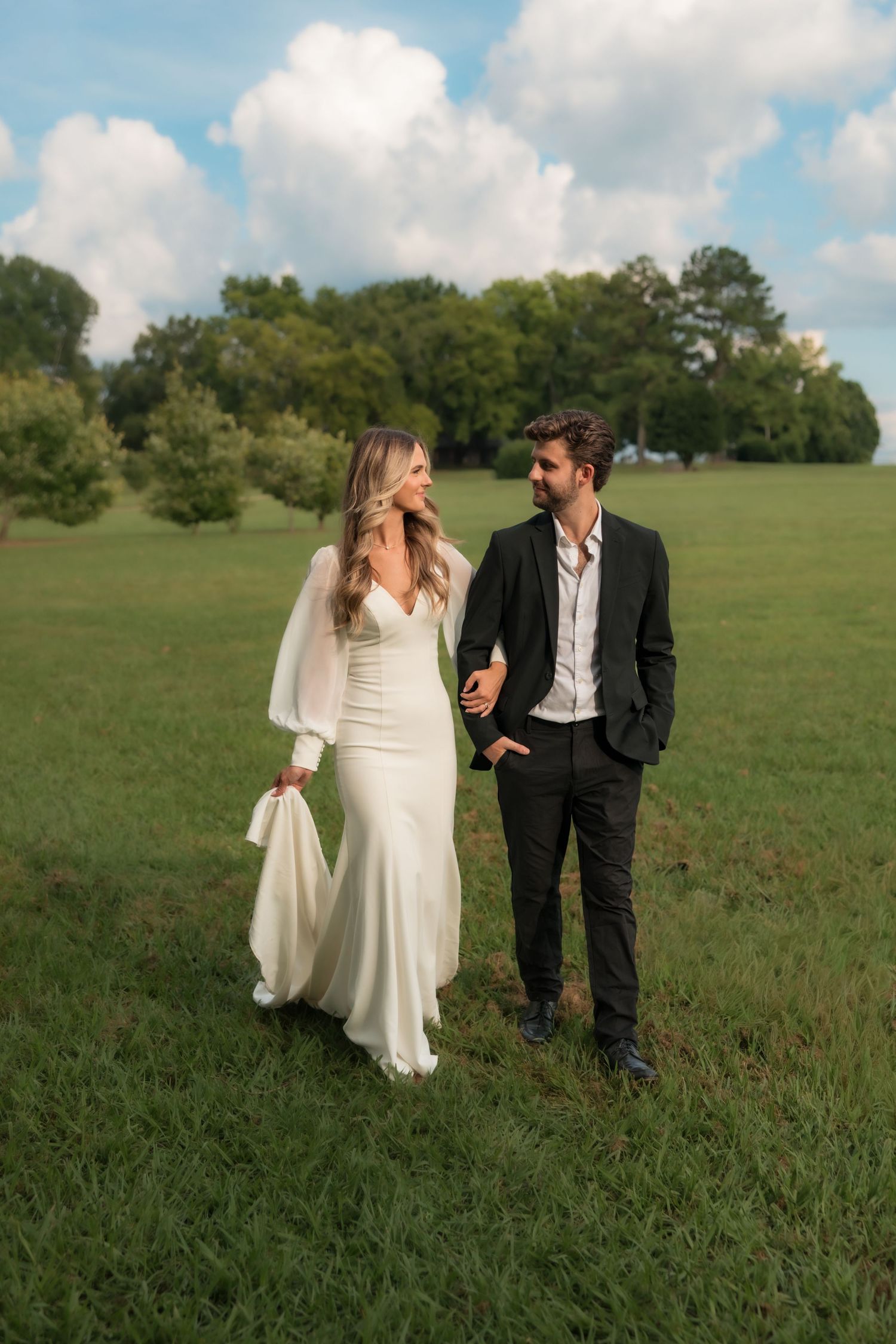 Bride and groom walk through lush green field, exchanging loving glances, bride in white gown with sheer sleeves.