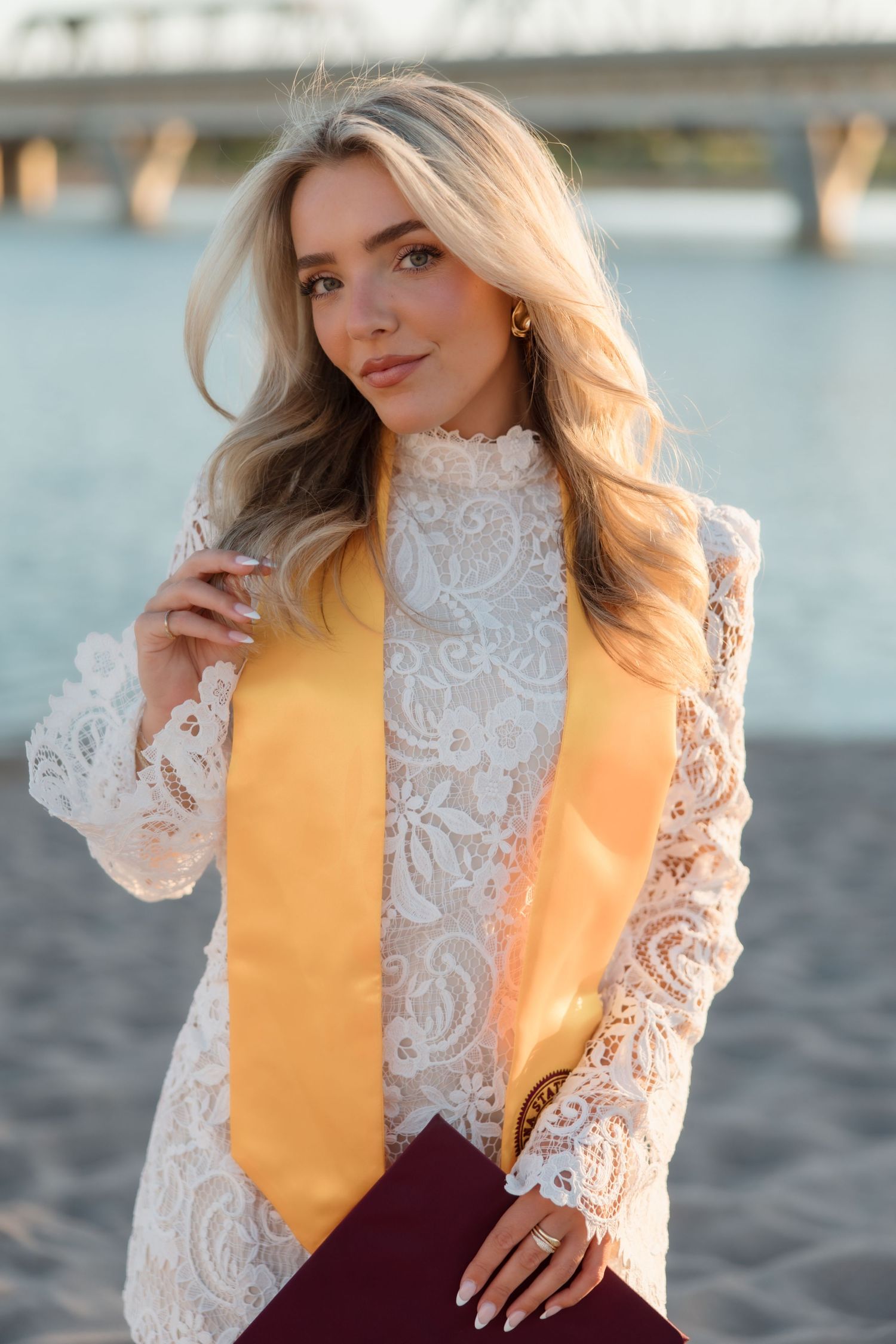 Blonde woman in white lace dress with gold graduation stole poses on sandy beach near bridge.