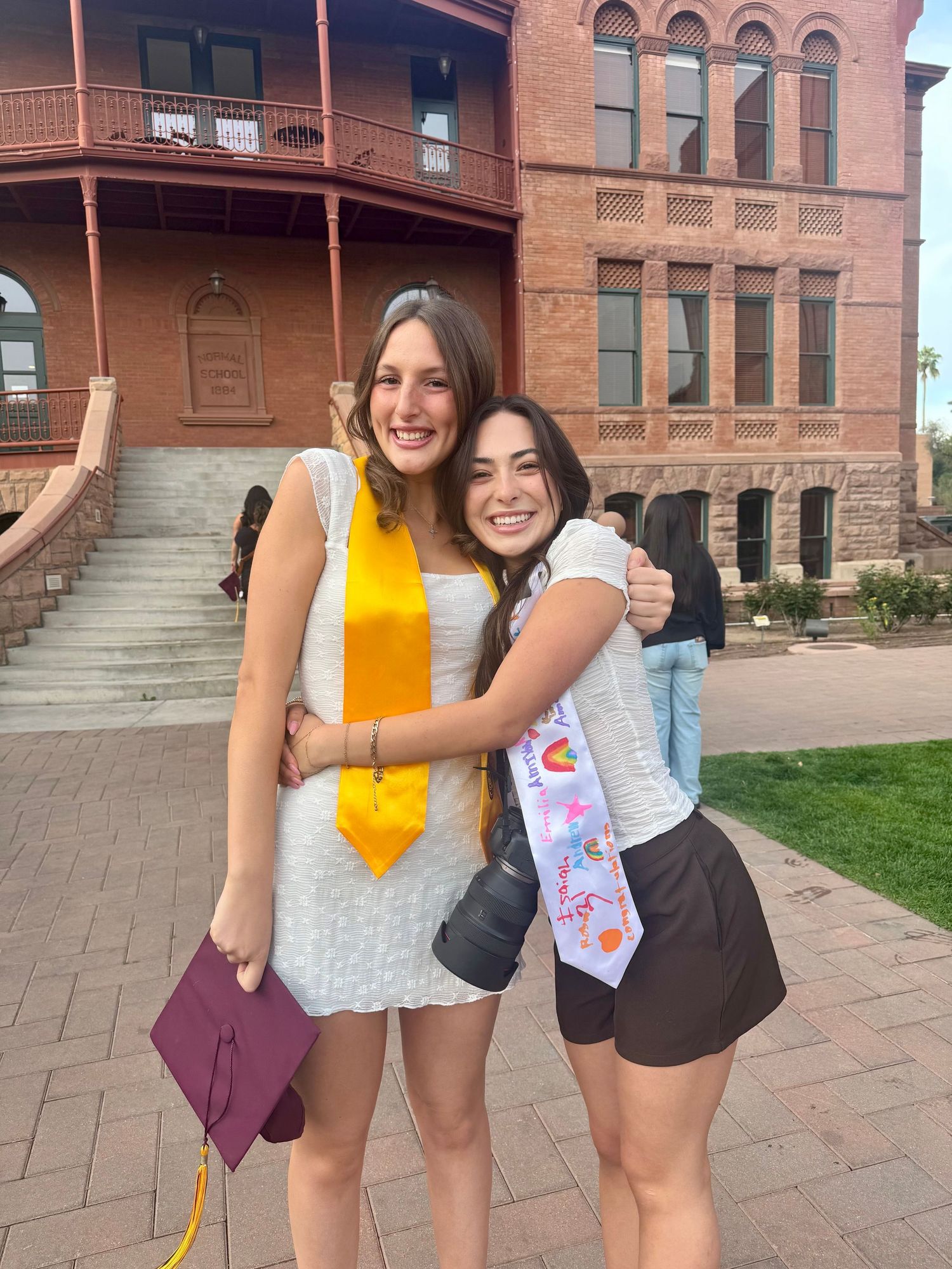 Two smiling young women hug outside historic brick building at college graduation celebration.
