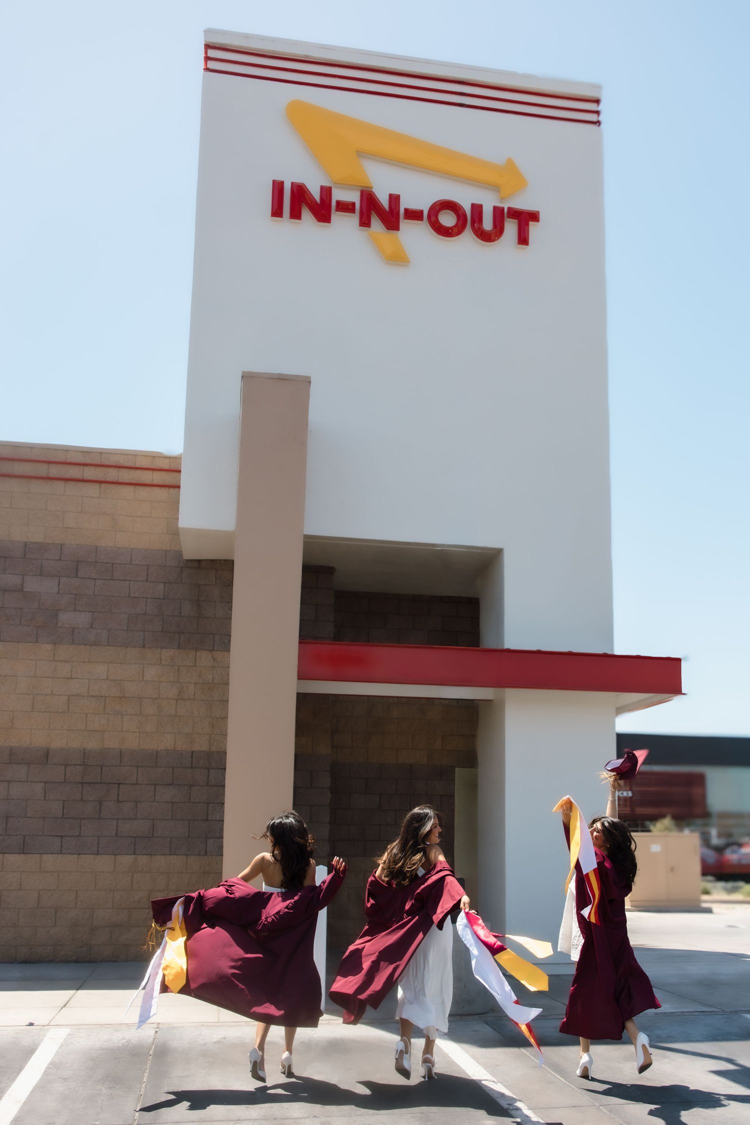 Three graduates in burgundy gowns celebrate outside an In-N-Out Burger, tossing their caps in the air.