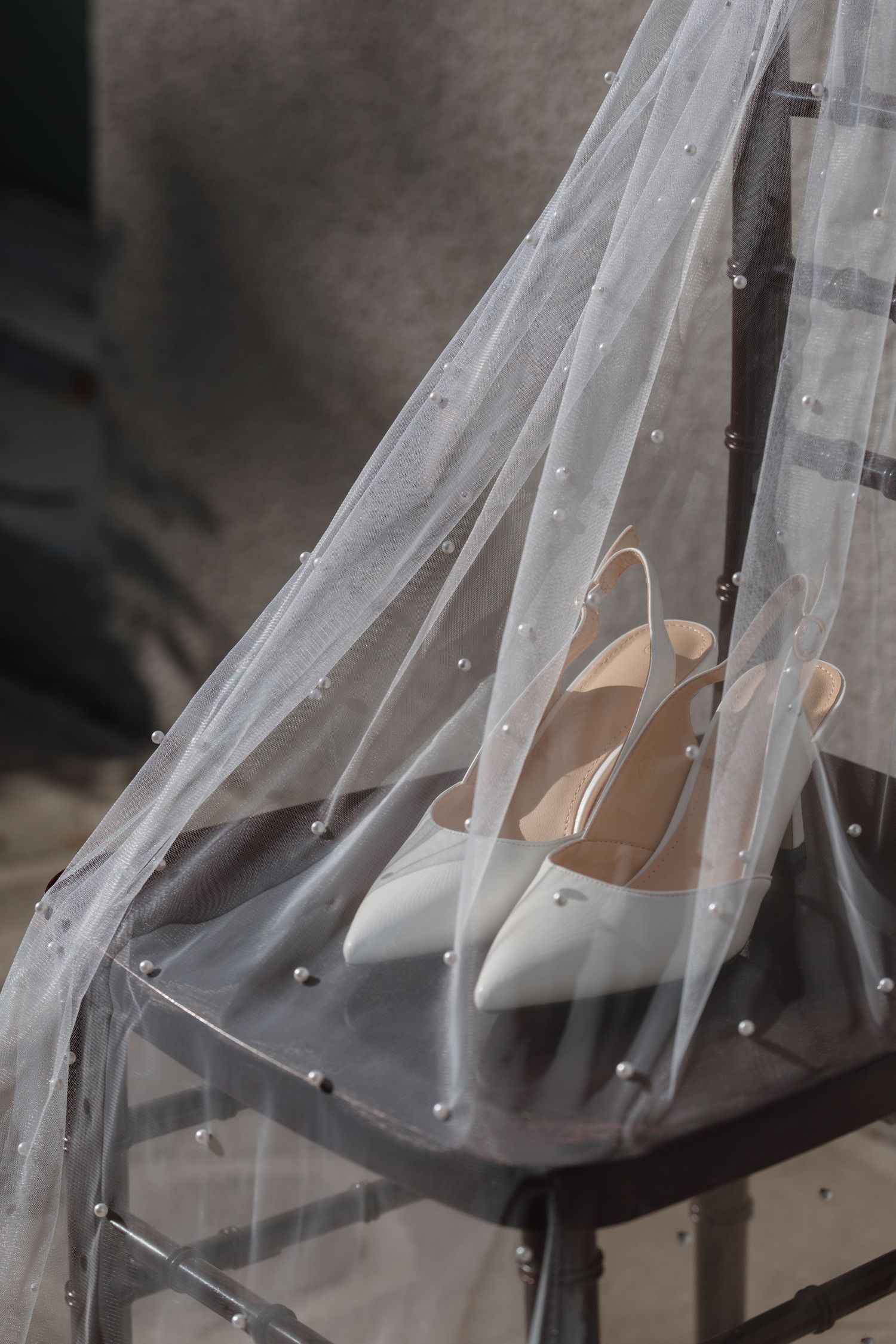 White slingback bridal heels resting on a chair draped with a pearl-embellished wedding veil.