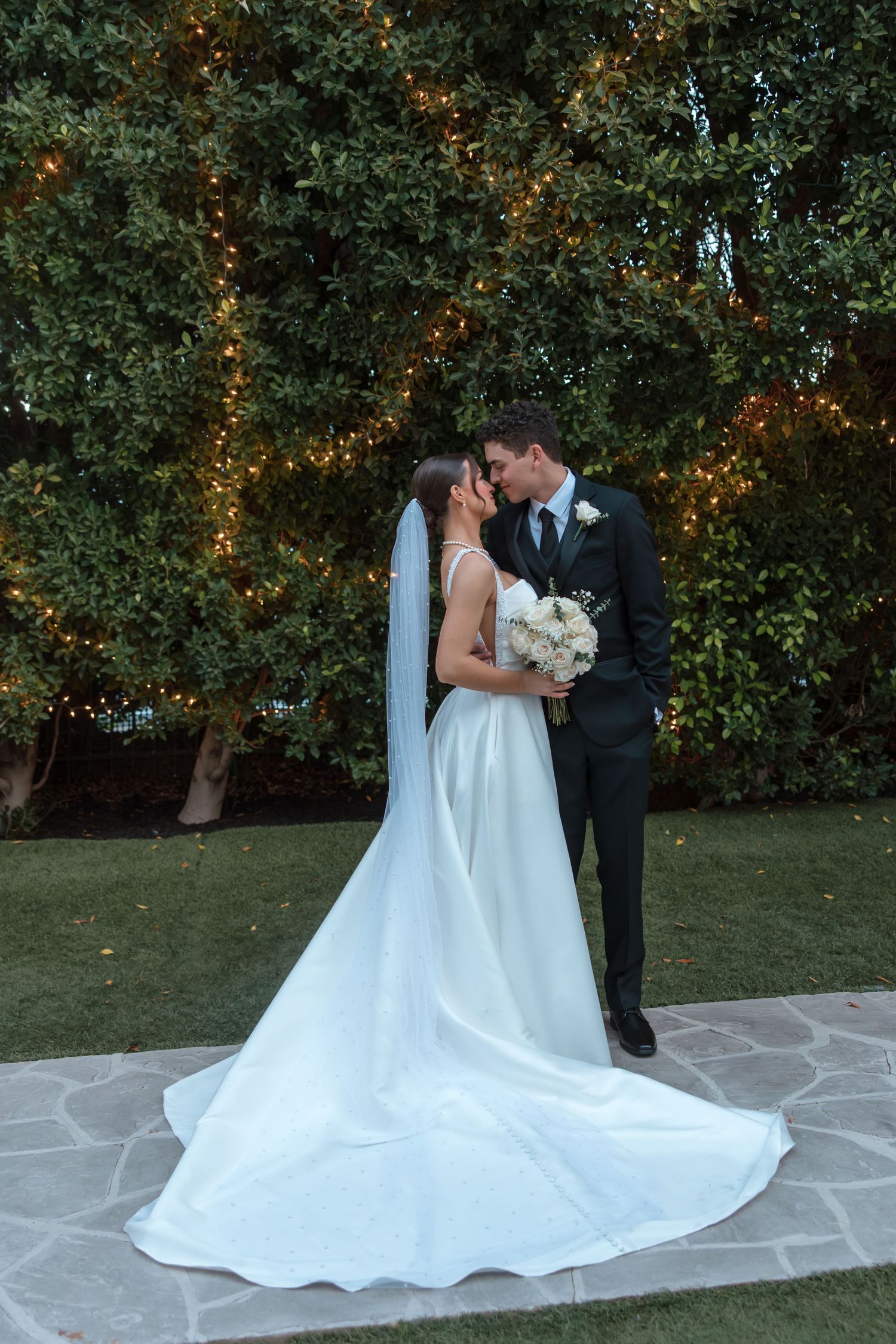 Bride and groom share a romantic kiss in front of a lighted hedge at their wedding.