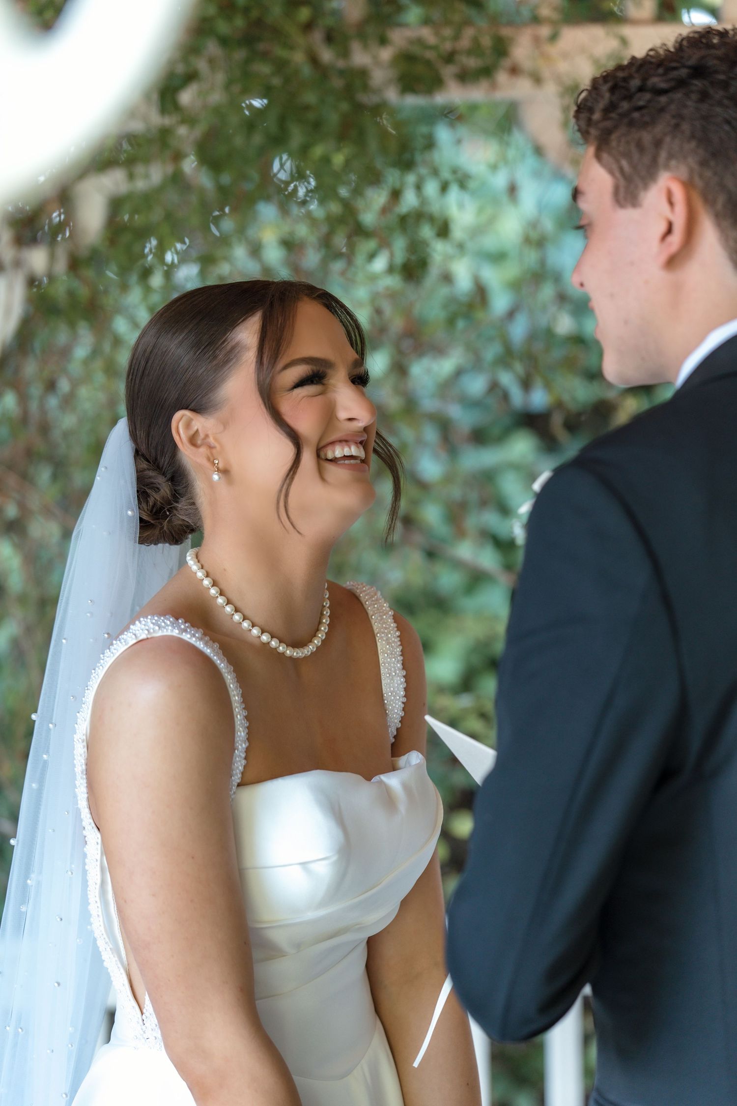 Joyful bride in pearl necklace and veil laughing during wedding ceremony with lush green backdrop.